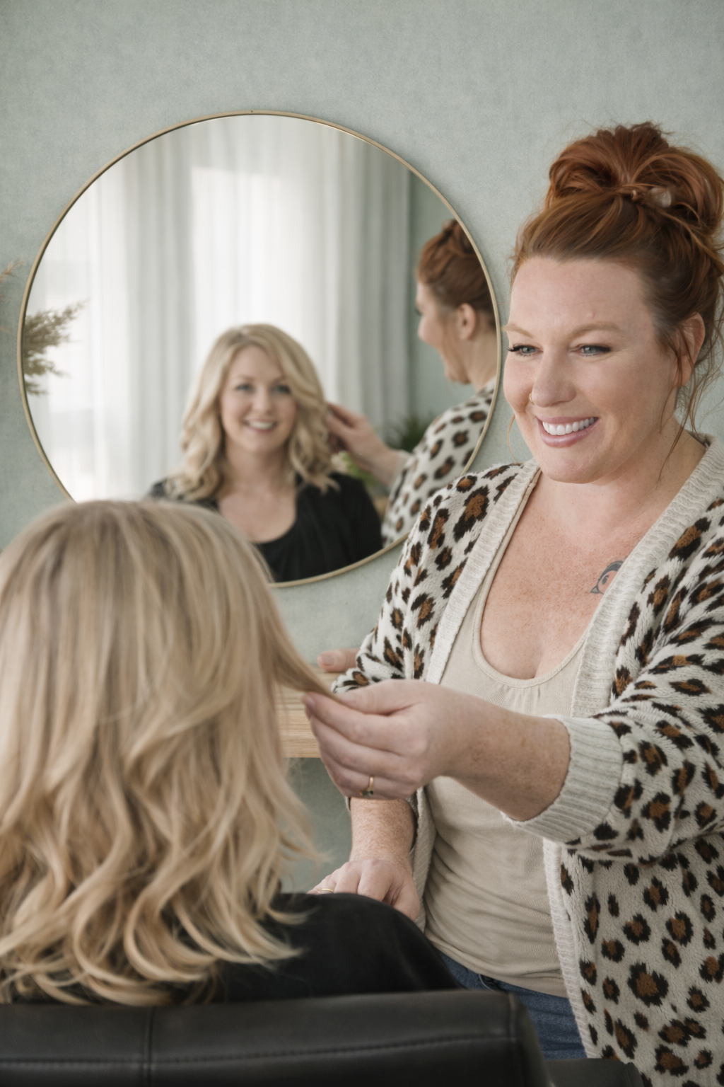 Ashley helping a woman with blonde hair in a salon, smiling as she styles her hair, with a mirror reflecting her face.