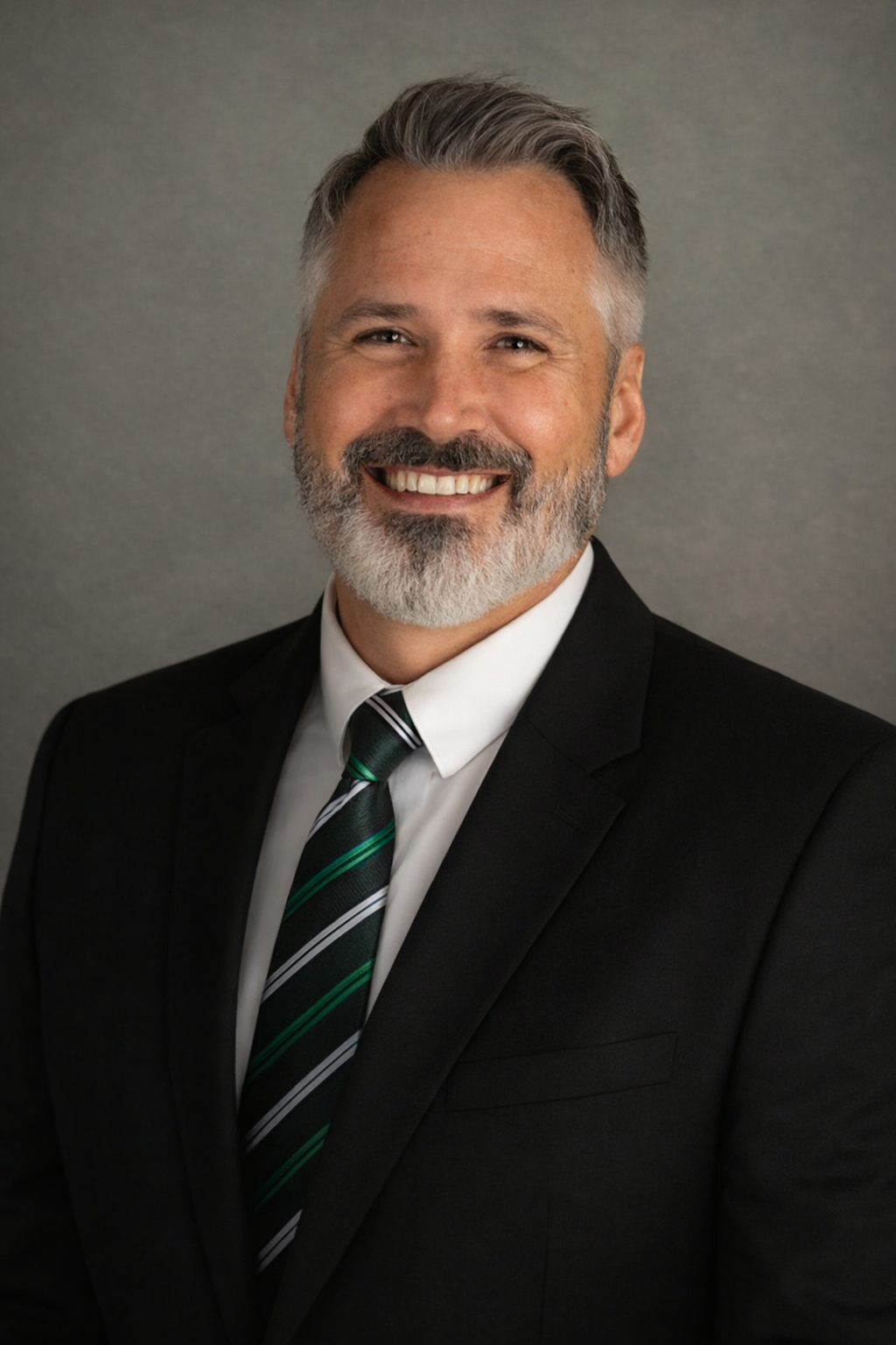 Headshot of a smiling man with gray hair and beard, wearing a black suit, white shirt, and a black, green, and white striped tie, against a plain gray background.