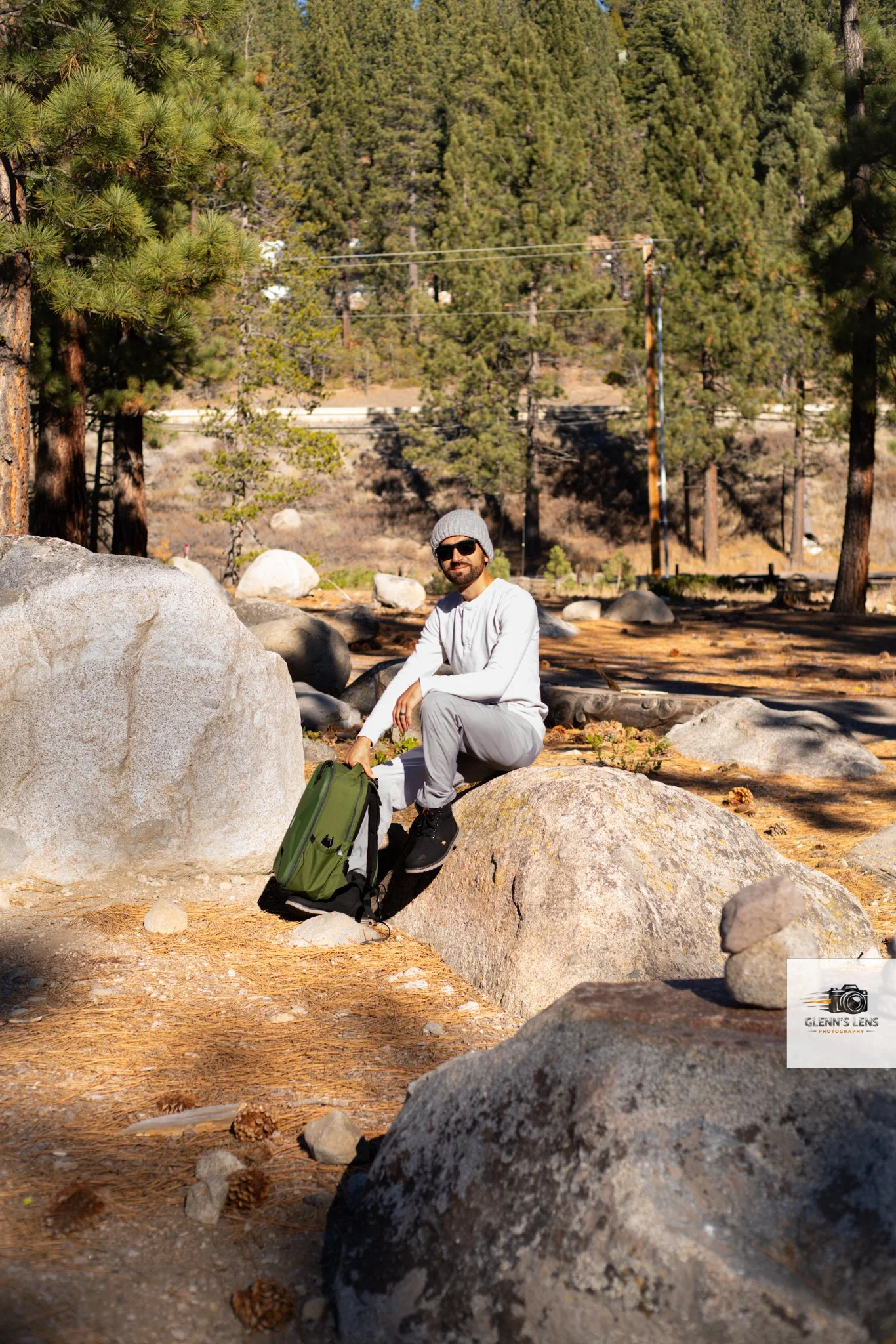 A man sitting on a large rock in a forested area, holding a green backpack, wearing sunglasses, a gray beanie, and light-colored hiking clothes, with tall pine trees and power lines in the background.
