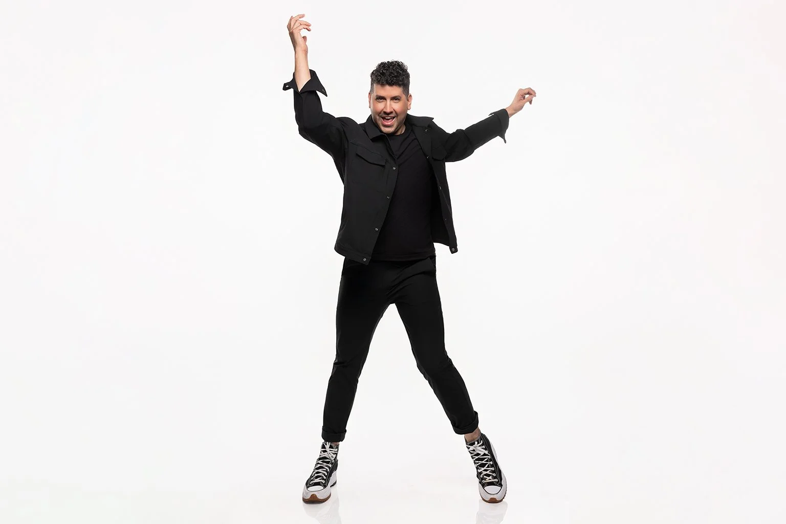A joyful man in black clothing and sneakers jumping and smiling against a white background.