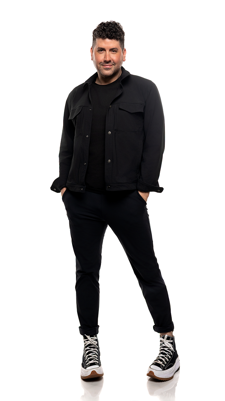 A man standing with his hands in his pockets, wearing a black jacket, black shirt, black pants, and high-top sneakers against a white background.
