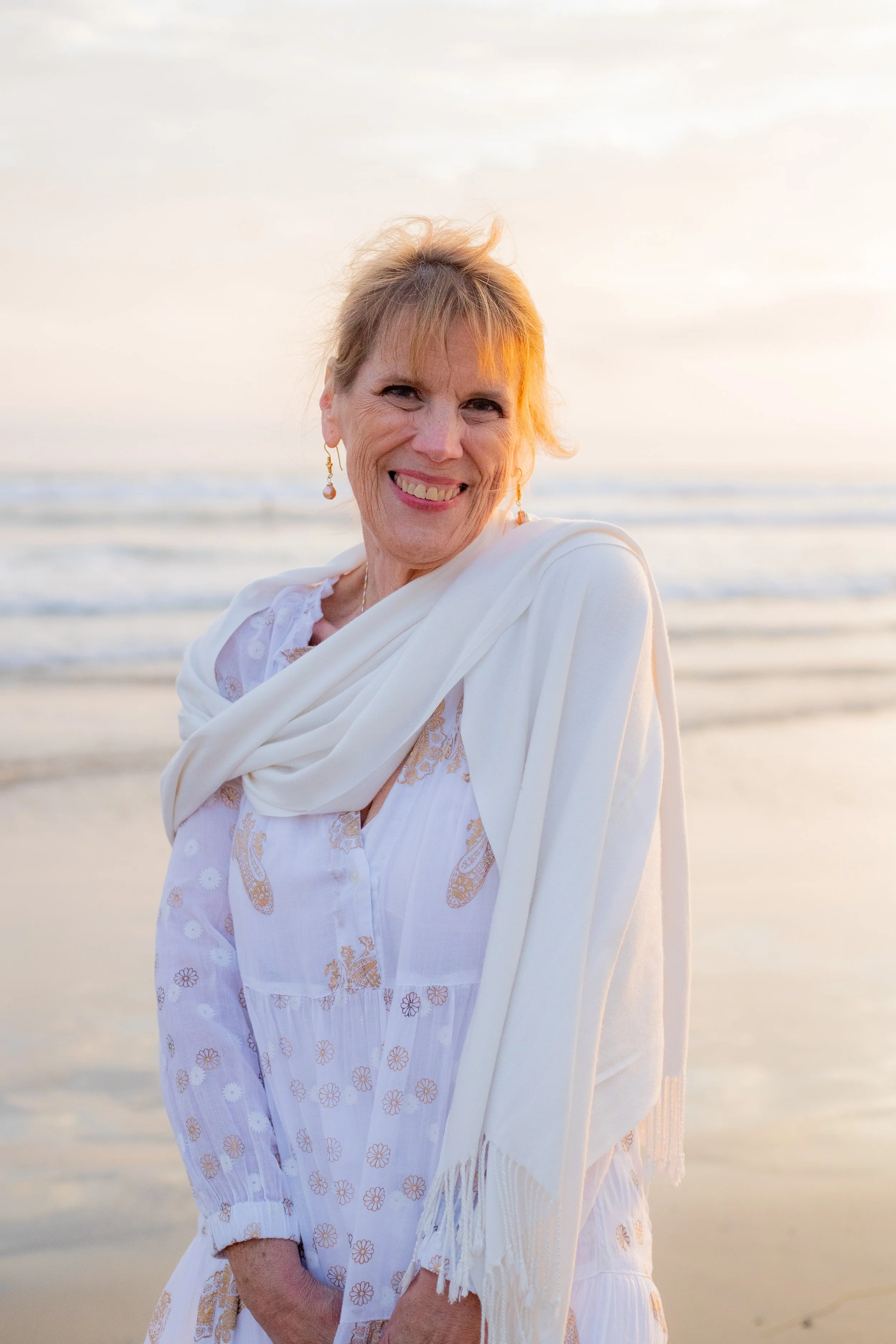 A smiling woman in a white dress and shawl standing on the beach at sunset.