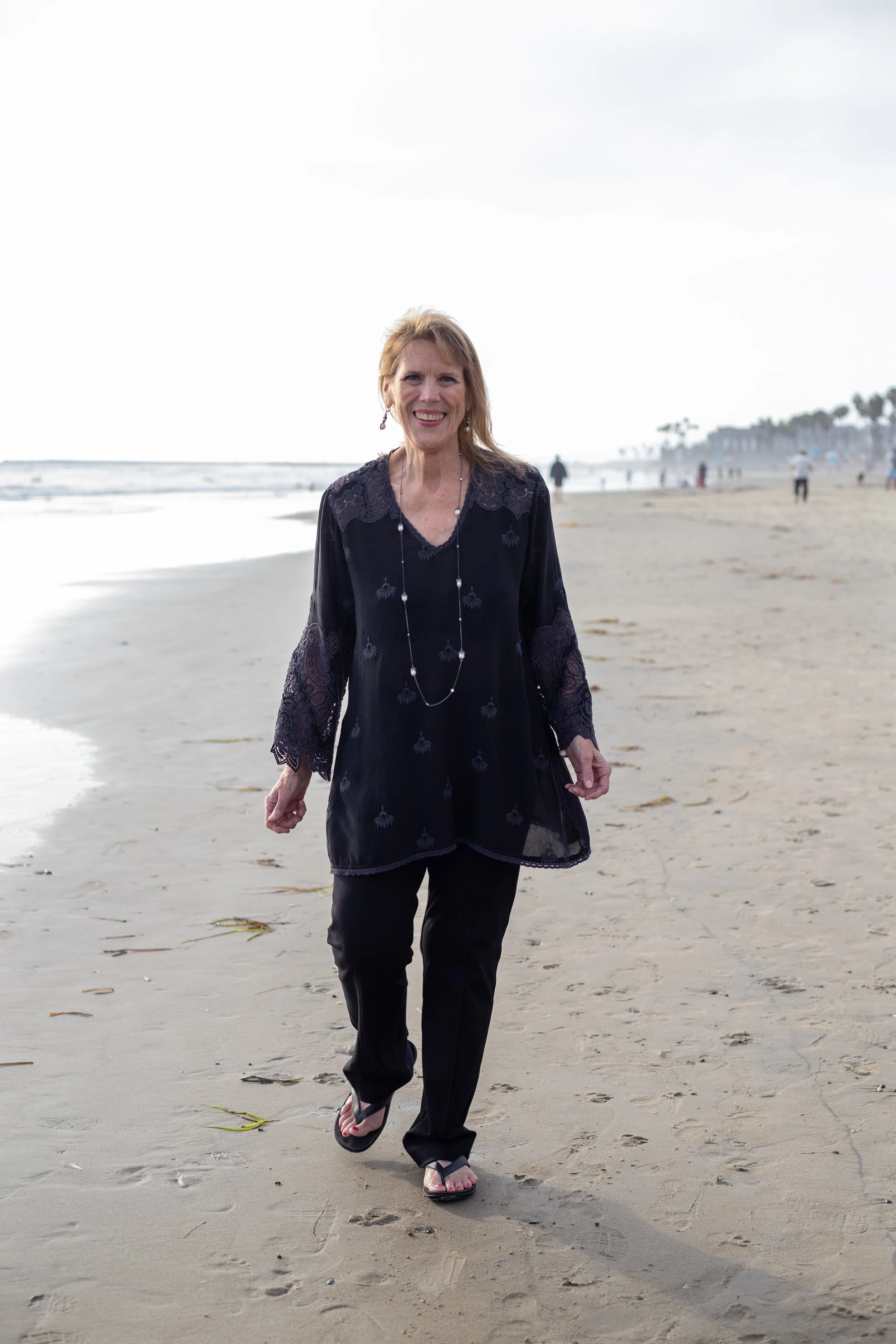Smiling woman with blond hair walking on the beach, wearing a black top and black pants, with ocean and cloudy sky in the background.