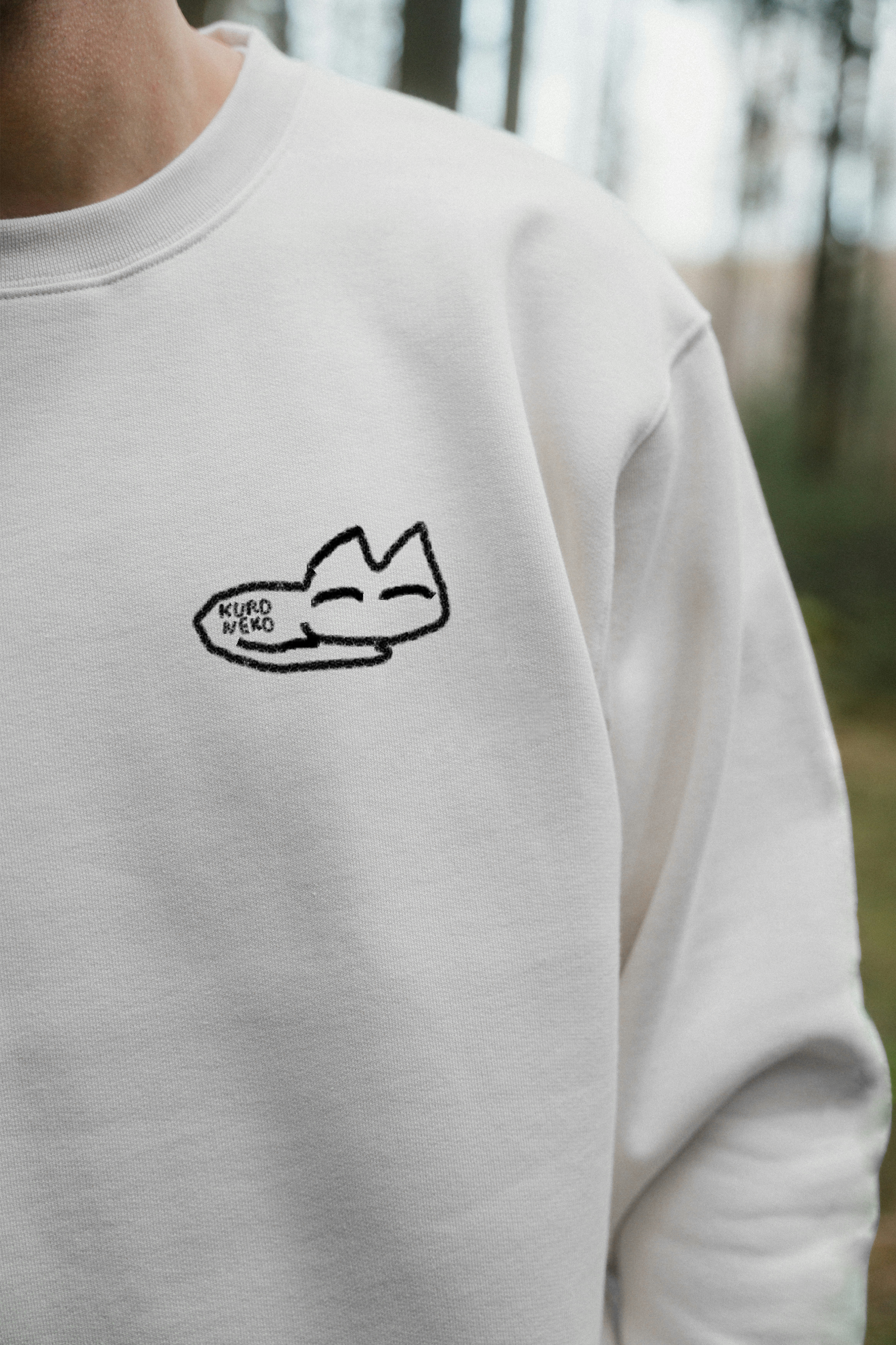 A person wearing a white sweatshirt with a black embroidered logo of a smiling cat and the words "KURO NEKO" on it, standing outdoors with trees in the background.