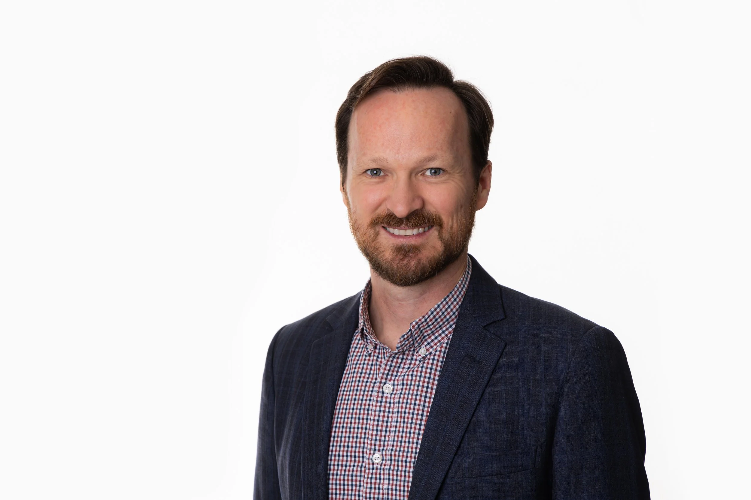 Headshot of a smiling man with a beard, wearing a dark blazer and a checkered shirt against a plain white background.