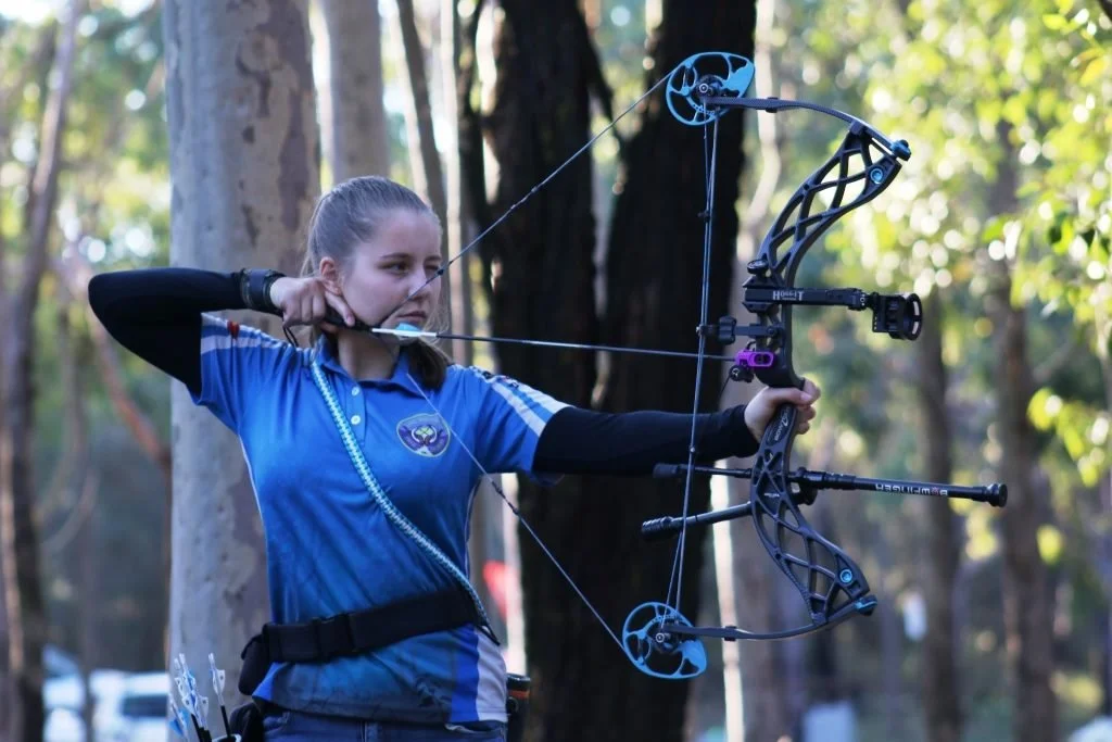 A woman in a blue sports shirt aims a modern compound bow with a camouflage pattern in an outdoor forested area.