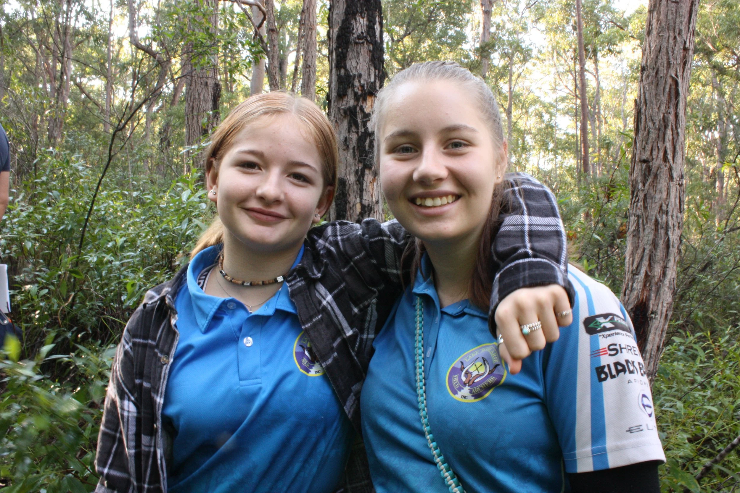 Two young women smiling and hugging in a wooded forest area, both wearing blue shirts with patches on the sleeve and chest.