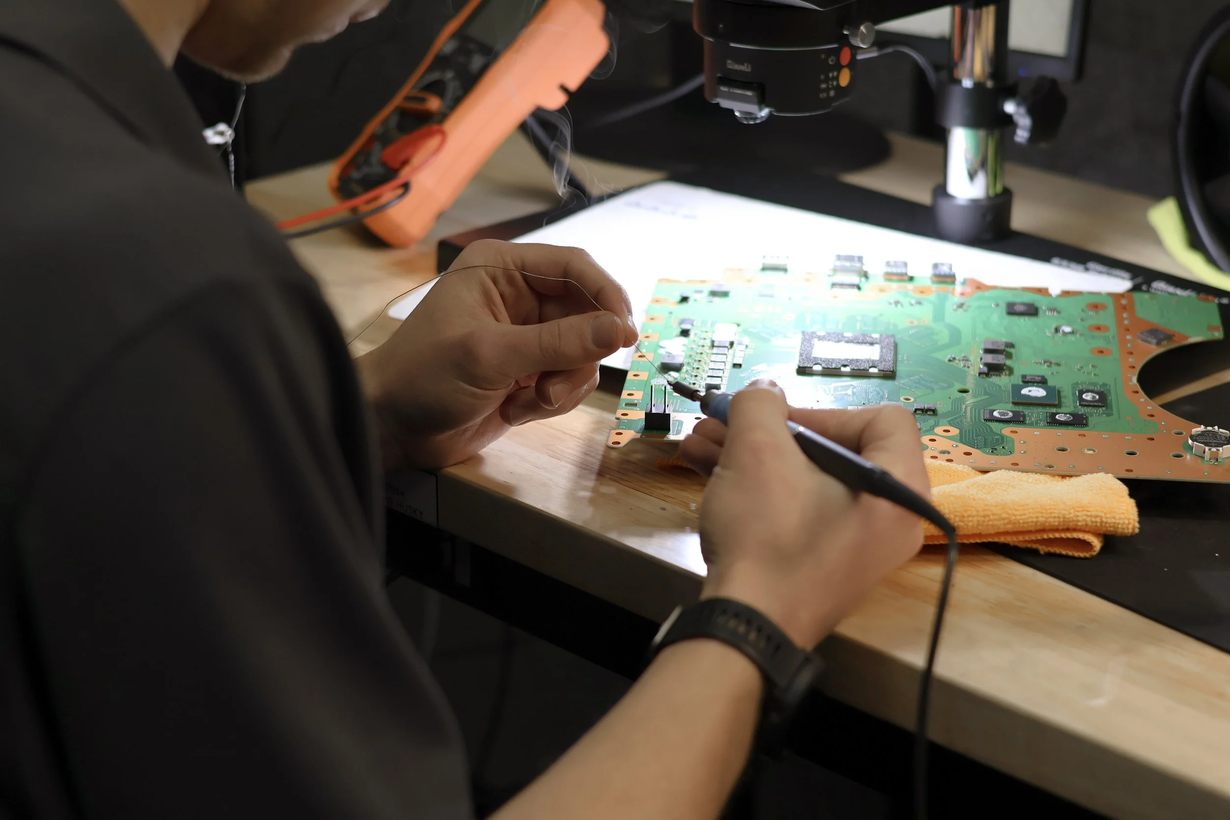 Person working on a green circuit board with tools, at a workbench with a lit surface and electronic equipment in the background.