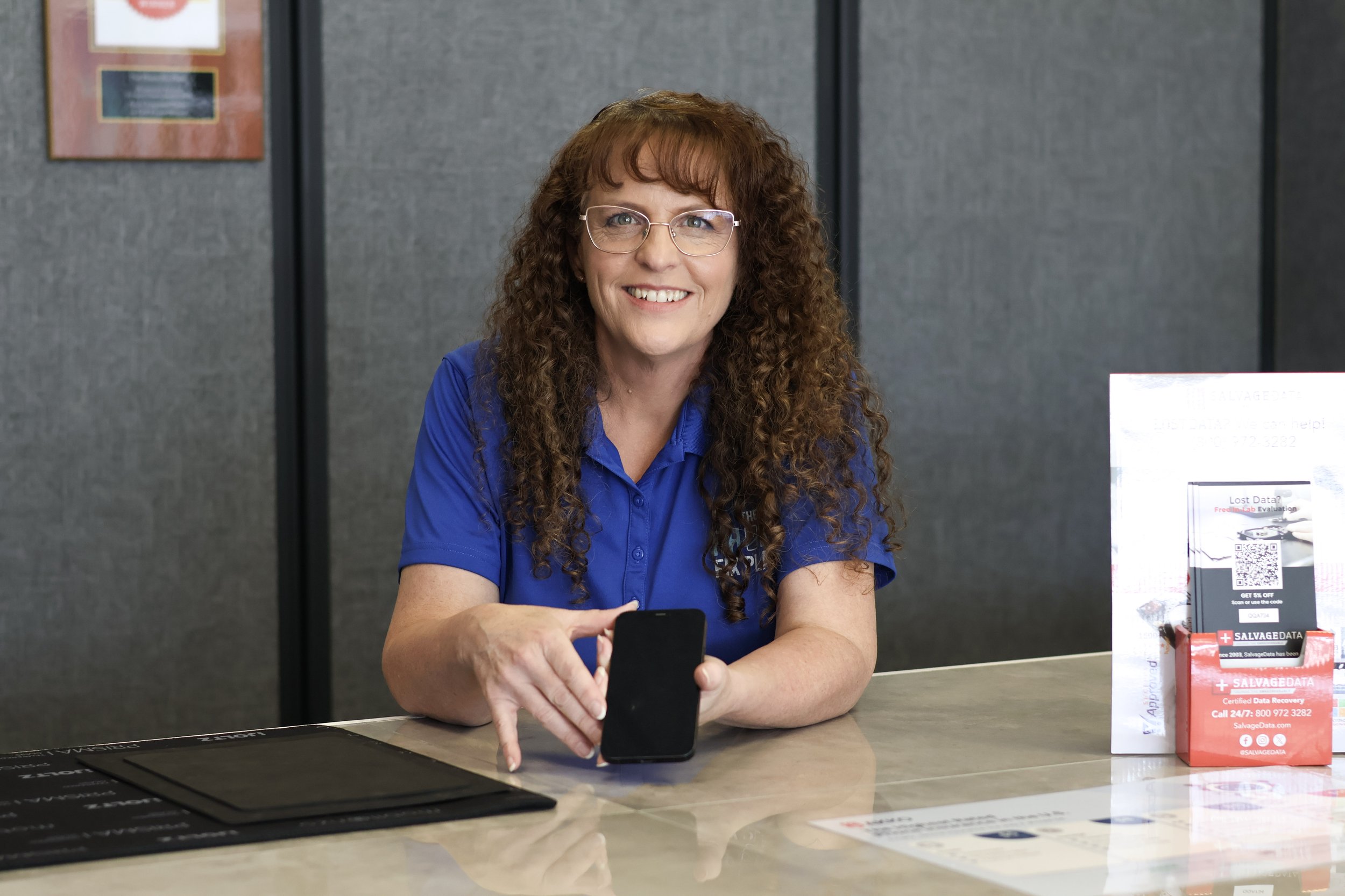 A woman with long curly hair, glasses, and a blue collared shirt sitting at a counter, holding a phone. There are informational signs on the counter and a gray partition behind her.