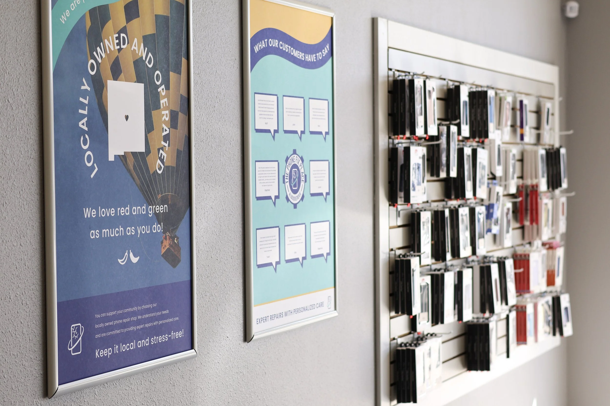 Display wall with posters and a magazine rack in a store or service center. The left poster promotes a locally owned and operated business supporting the community, emphasizing keeping it local and stress-free. The middle poster highlights customer feedback with speech bubbles and the logo of The Phone Fix. The right side features a white slatwall with various phone cases and accessories displayed.