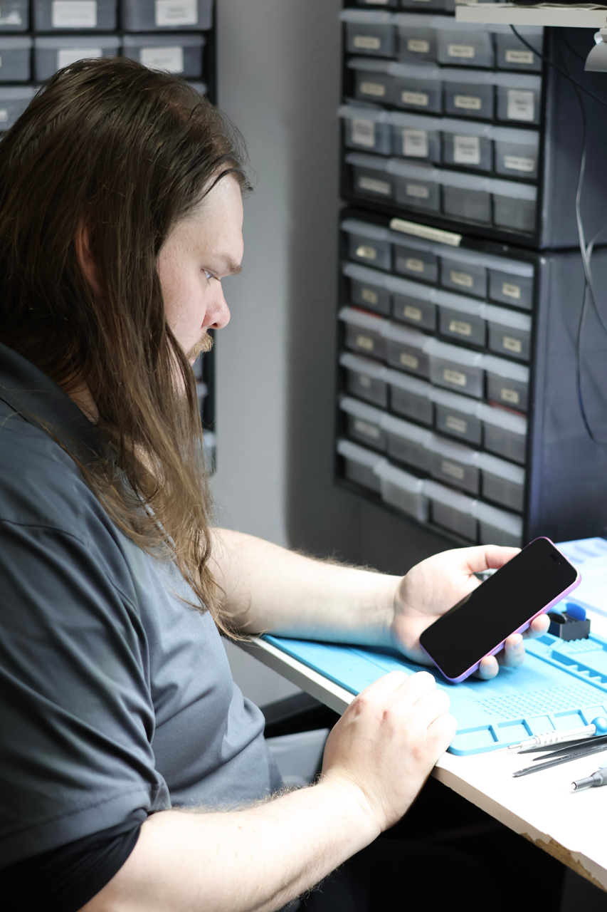 A man with long brown hair and a gray shirt sitting at a workbench, looking at his smartphone in a workshop or repair space with organized drawers in the background.