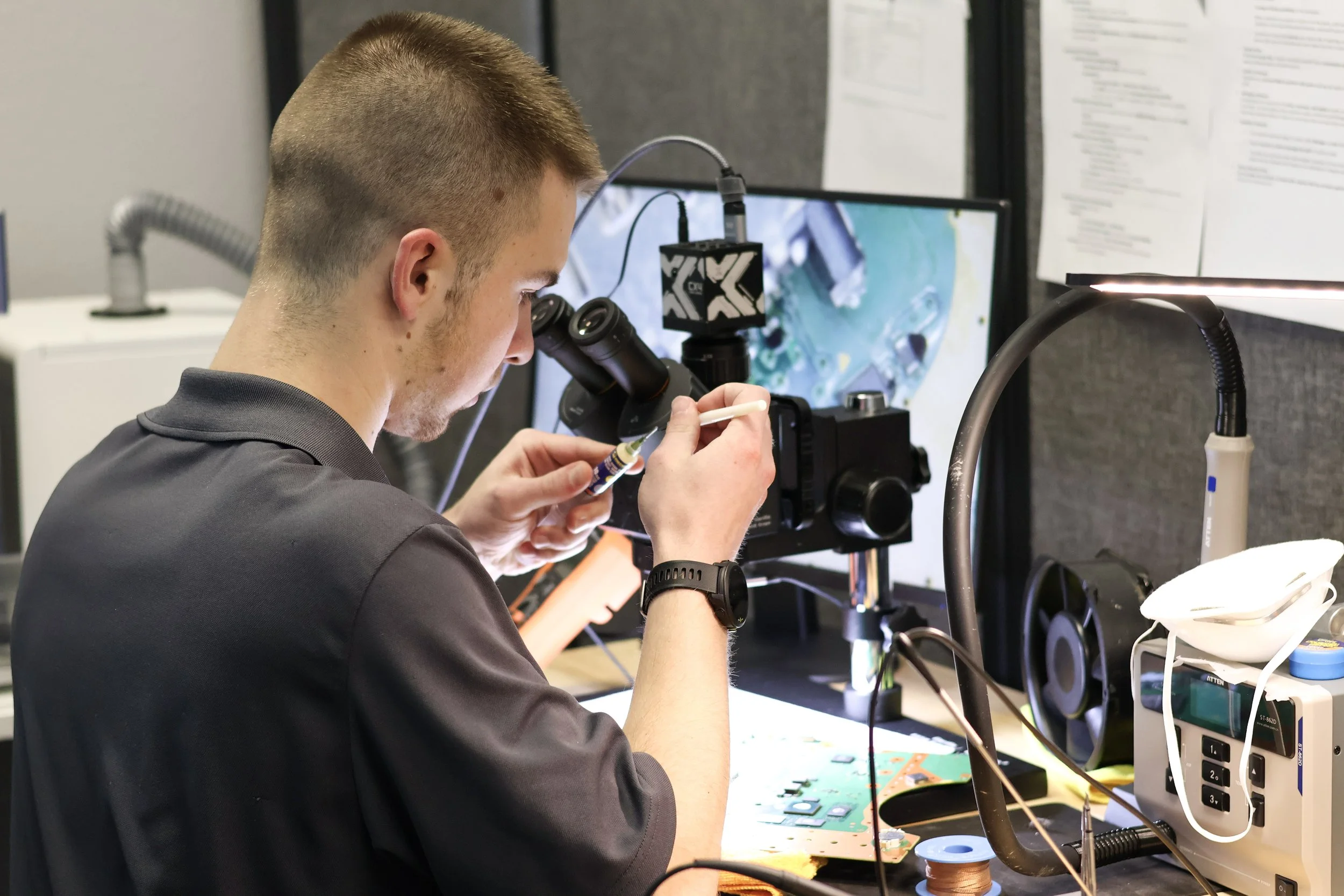 A young man working with electronic components at a workspace, using a microscope and soldering tools.