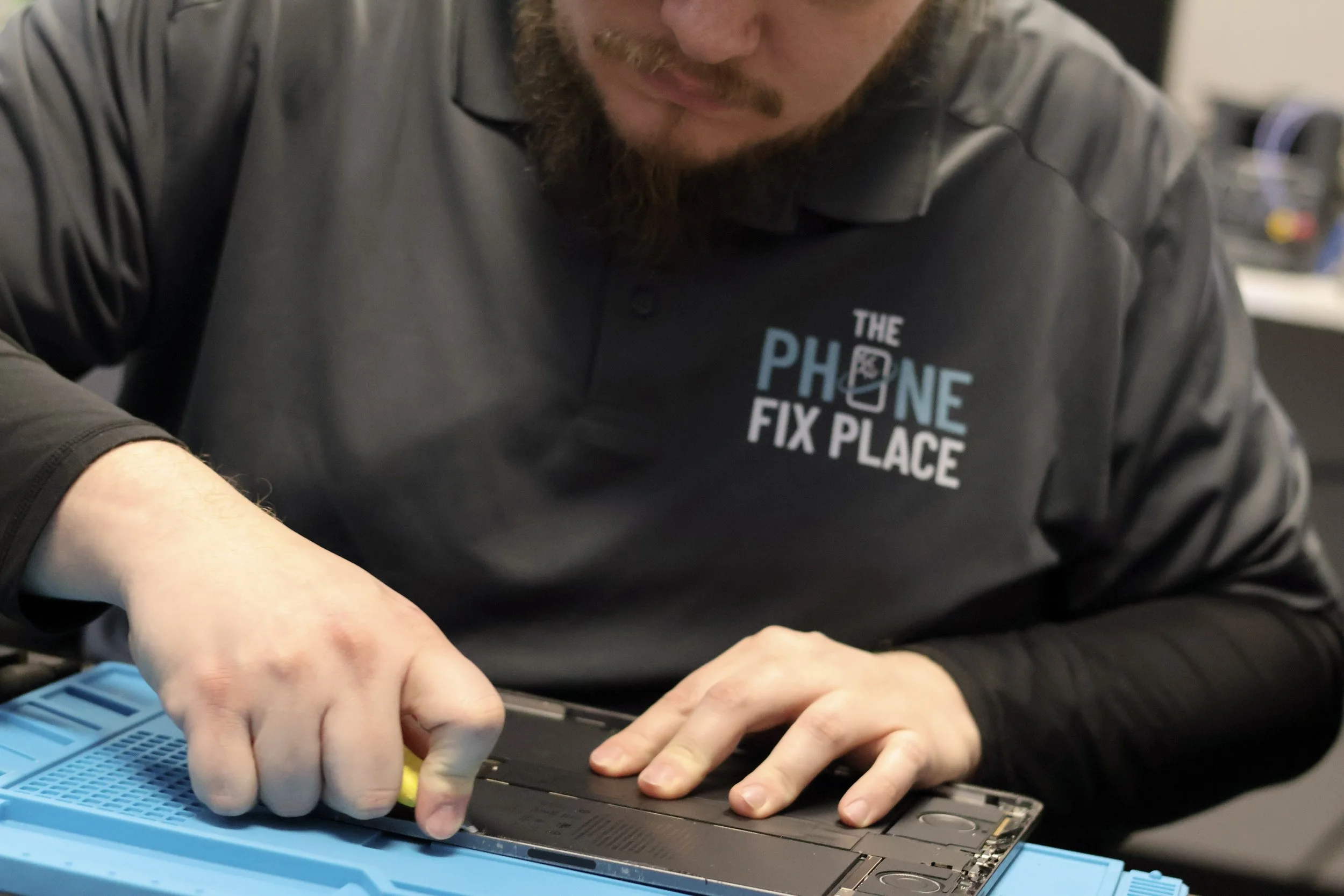 A technician working on a phone repair at a repair shop. He is wearing a black shirt with the logo 'The Phone Fix Place' and is using a tool to work on a disassembled phone placed on a blue work surface.