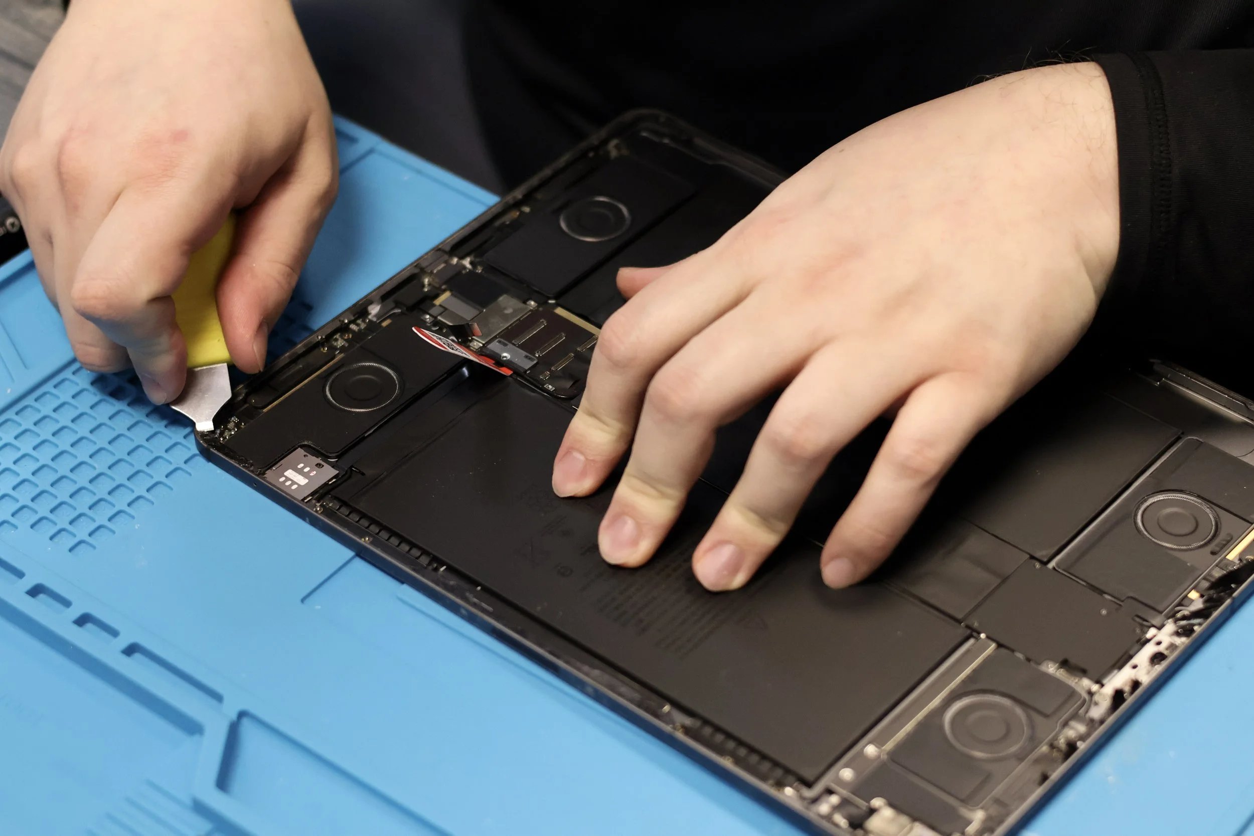 A person repairing a disassembled laptop, using a yellow plastic tool to work on the internal components, with a blue anti-static mat underneath.
