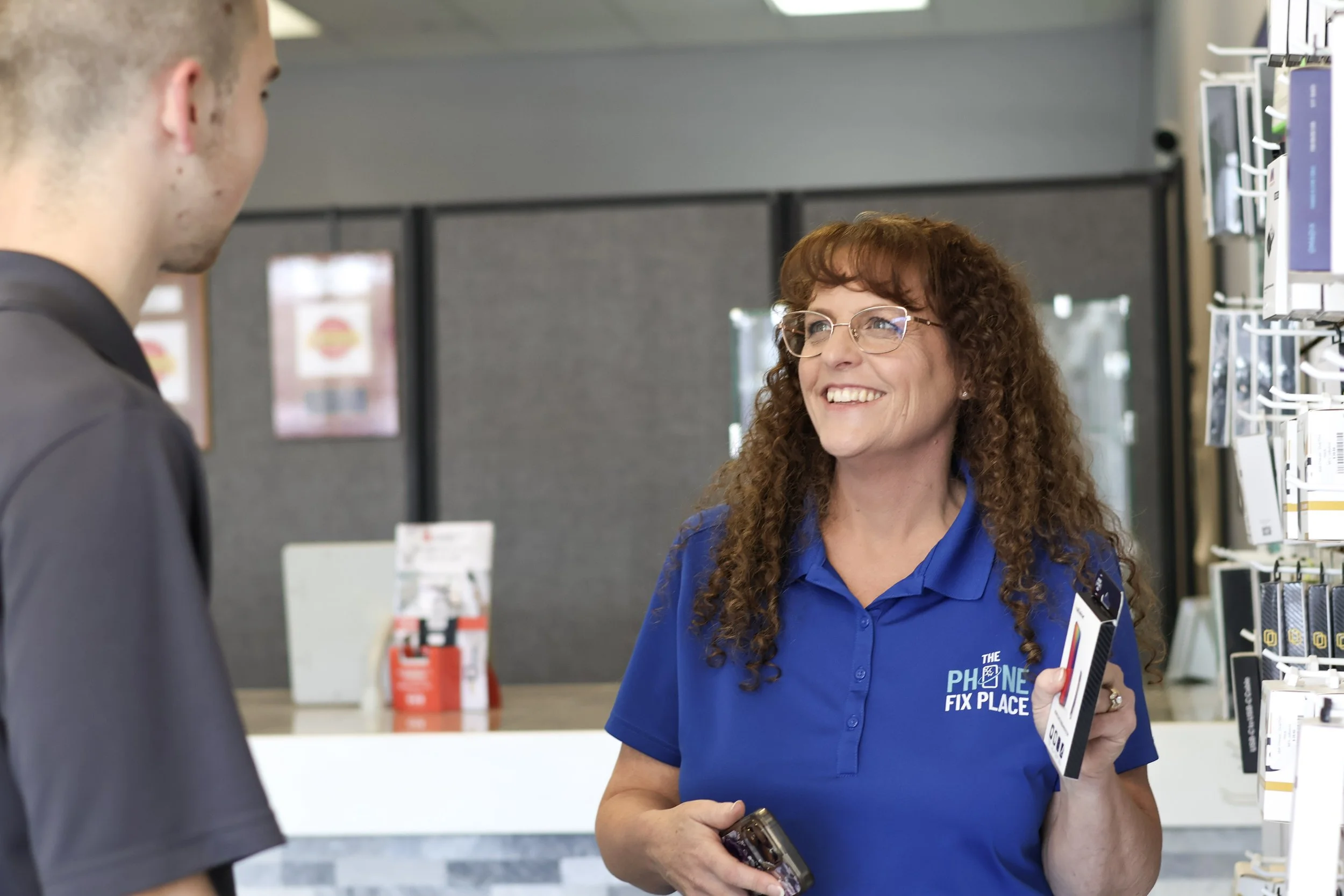 A smiling woman at a phone store wearing a blue polo shirt with a logo, talking to a man with a phone in her hand.