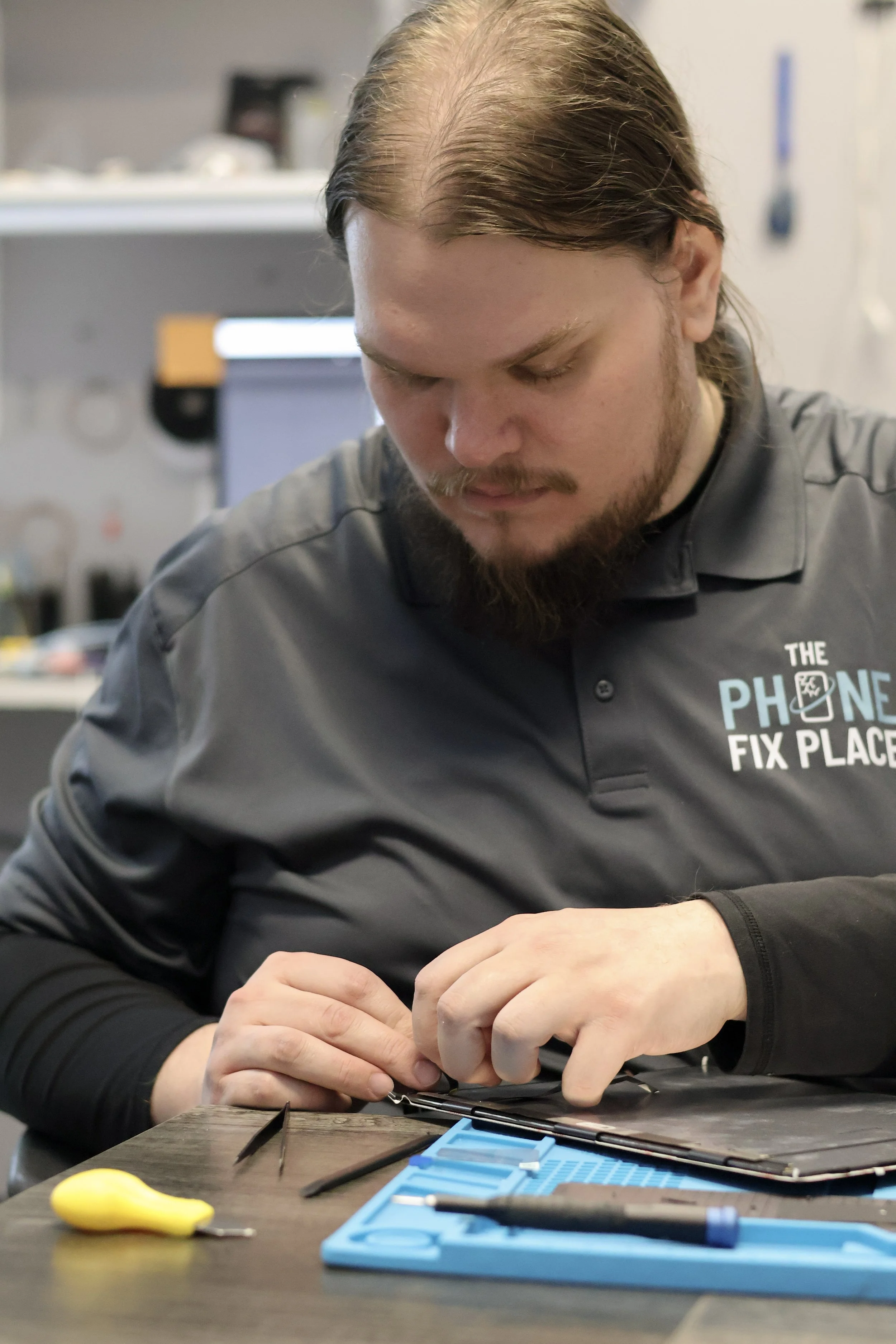 A man working on a phone repair at a workbench, tools and a yellow screwdriver nearby, wearing a gray shirt that reads 'The Phone Fix Place'.