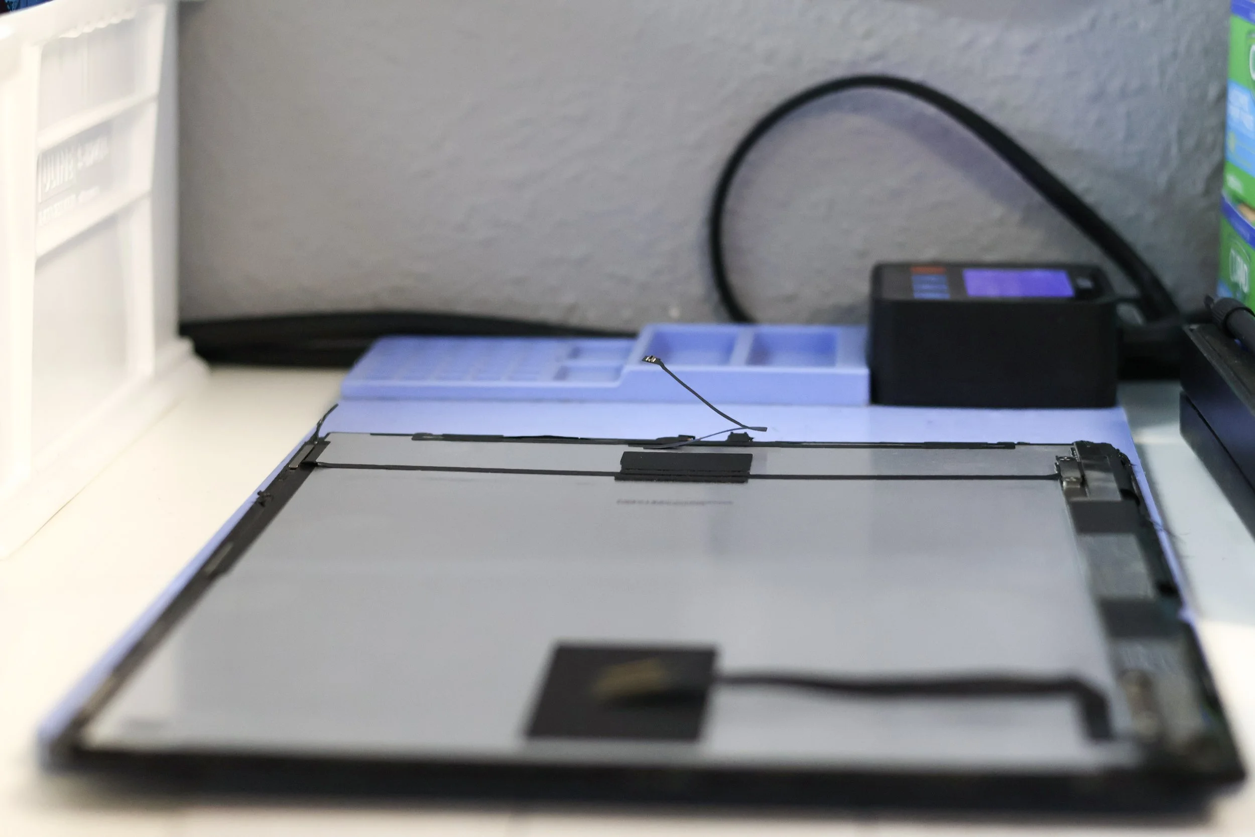 Disassembled tablet or laptop screen with internal components exposed on a desk, with a purple tray, a charger, and a box of printer ink nearby.