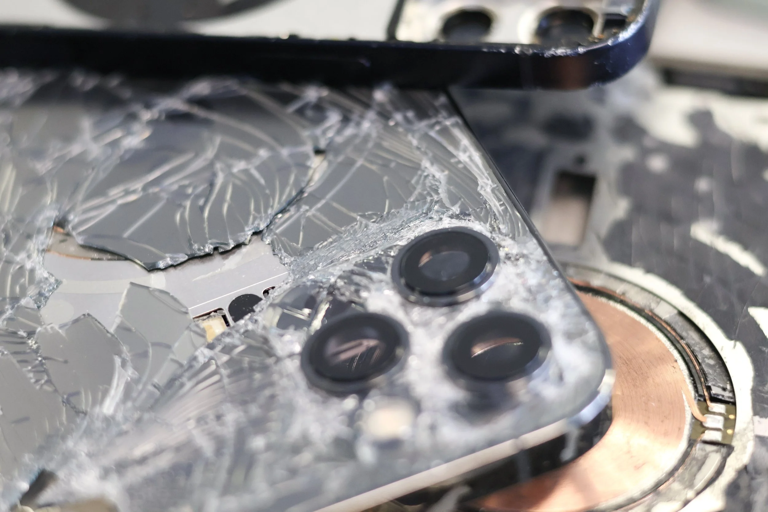 Close-up of a smartphone with a shattered screen, showing cracks radiating from the top left corner near the camera module, resting on a circular copper-colored component.