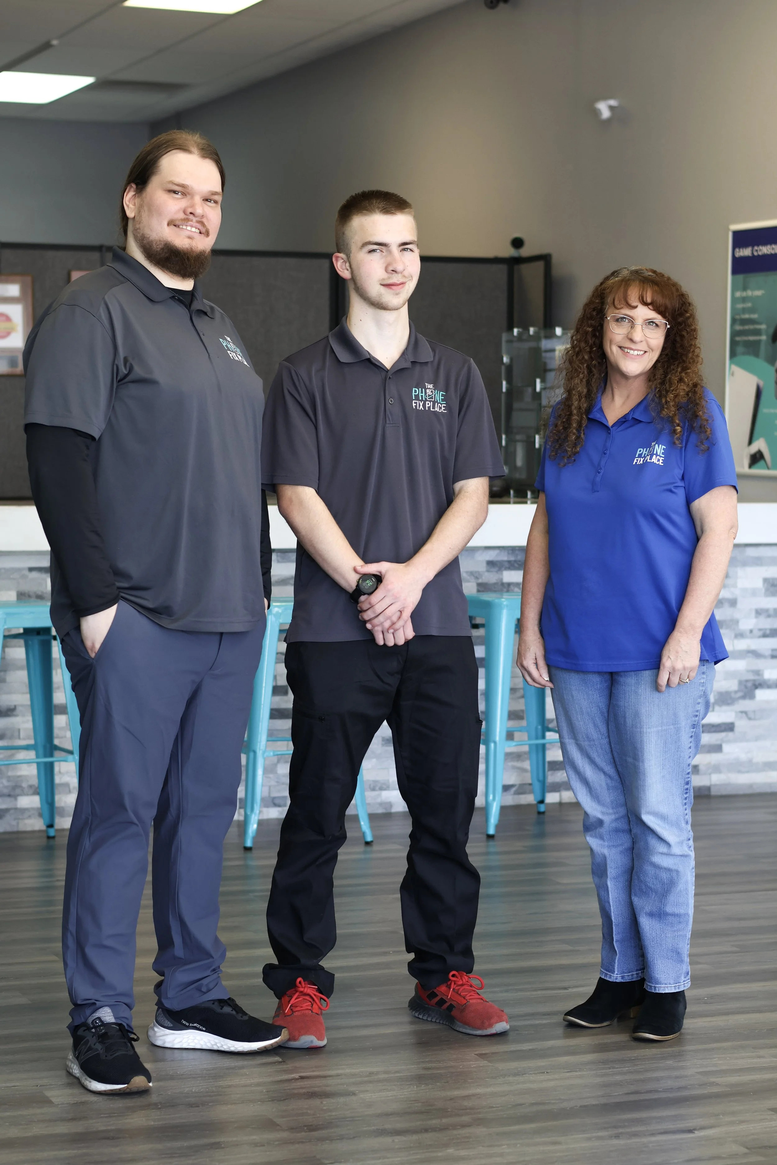 Three employees standing inside a modern office or lobby area, smiling at the camera. Two men and one woman, all wearing branded polo shirts with the company logo 'The Phone Fix Place', are in front of a bar-height counter with turquoise stools and a patterned backsplash.