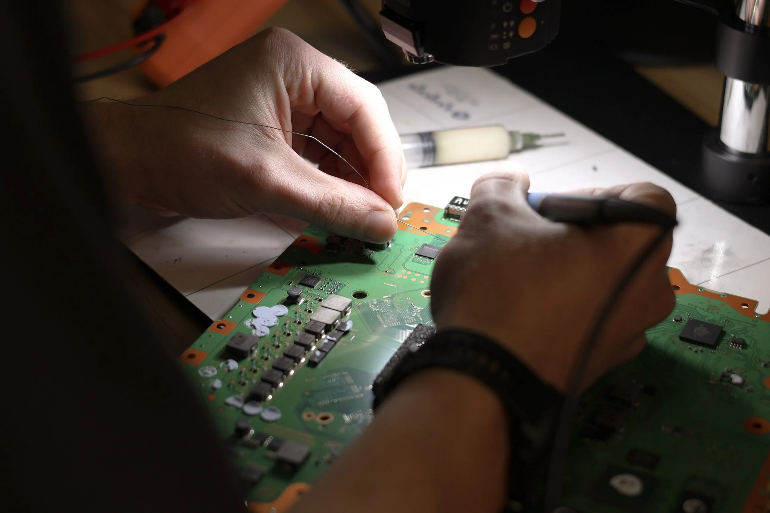 Person working on electronic circuit board, using a tool with wires attached, in a workshop setting.