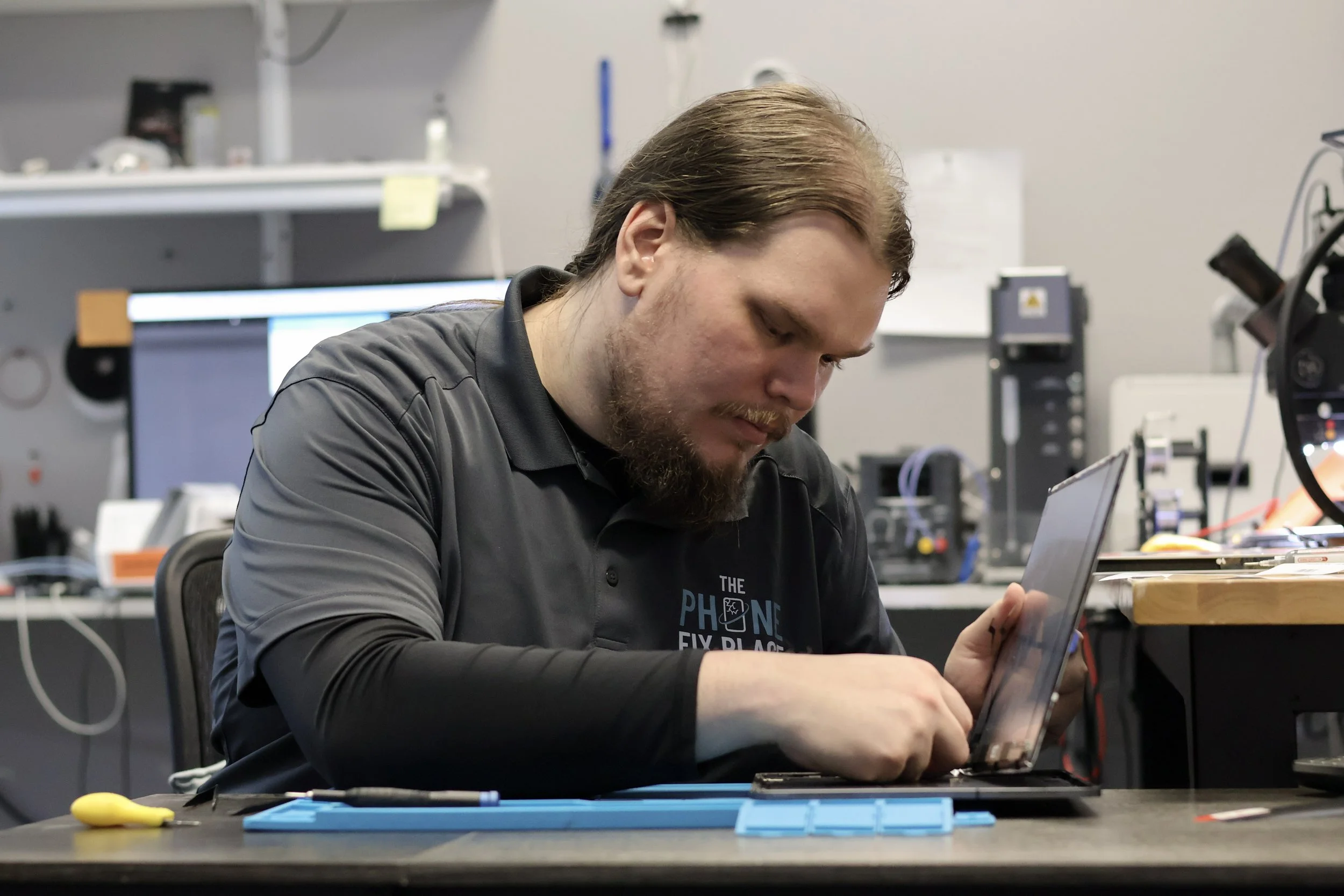 A man working on a laptop in a workshop or laboratory setting, with tools and electronic equipment on the table and shelves in the background.