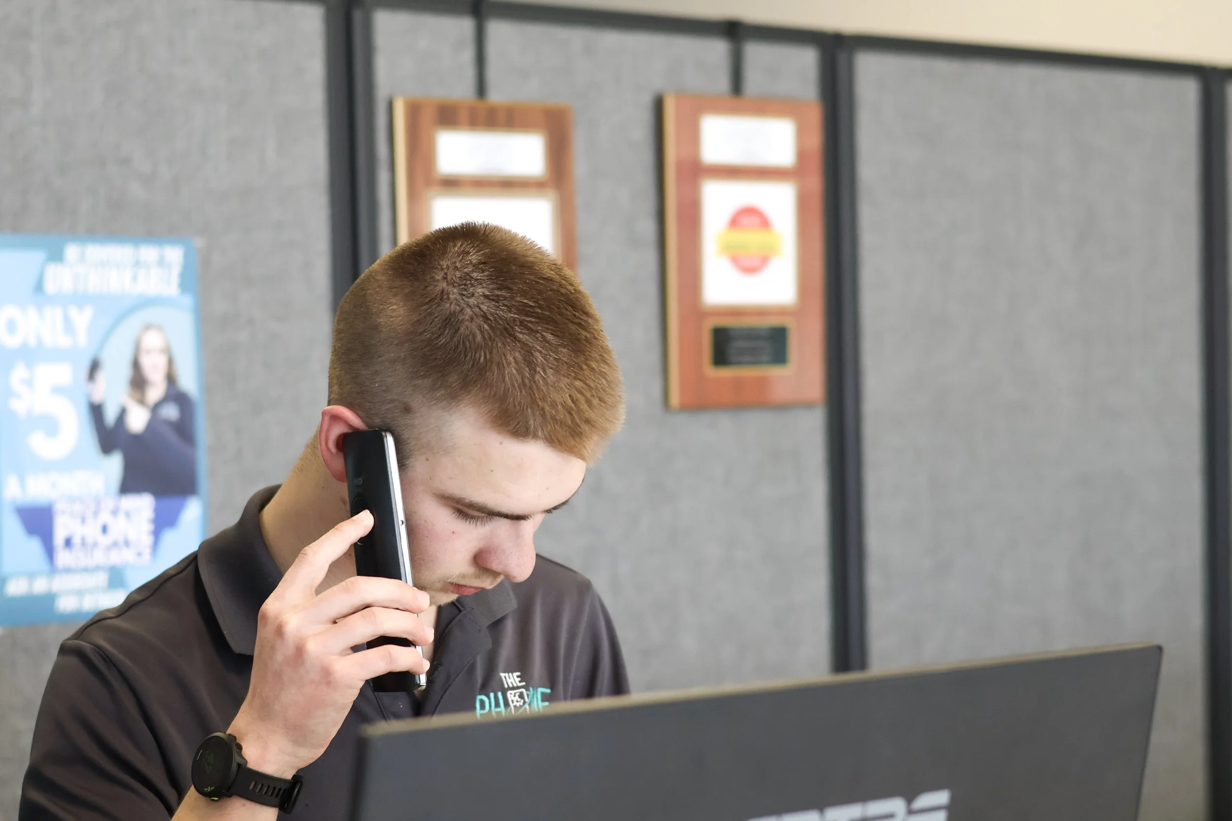 Young man talking on a phone and working on a laptop at a desk in an indoor setting.