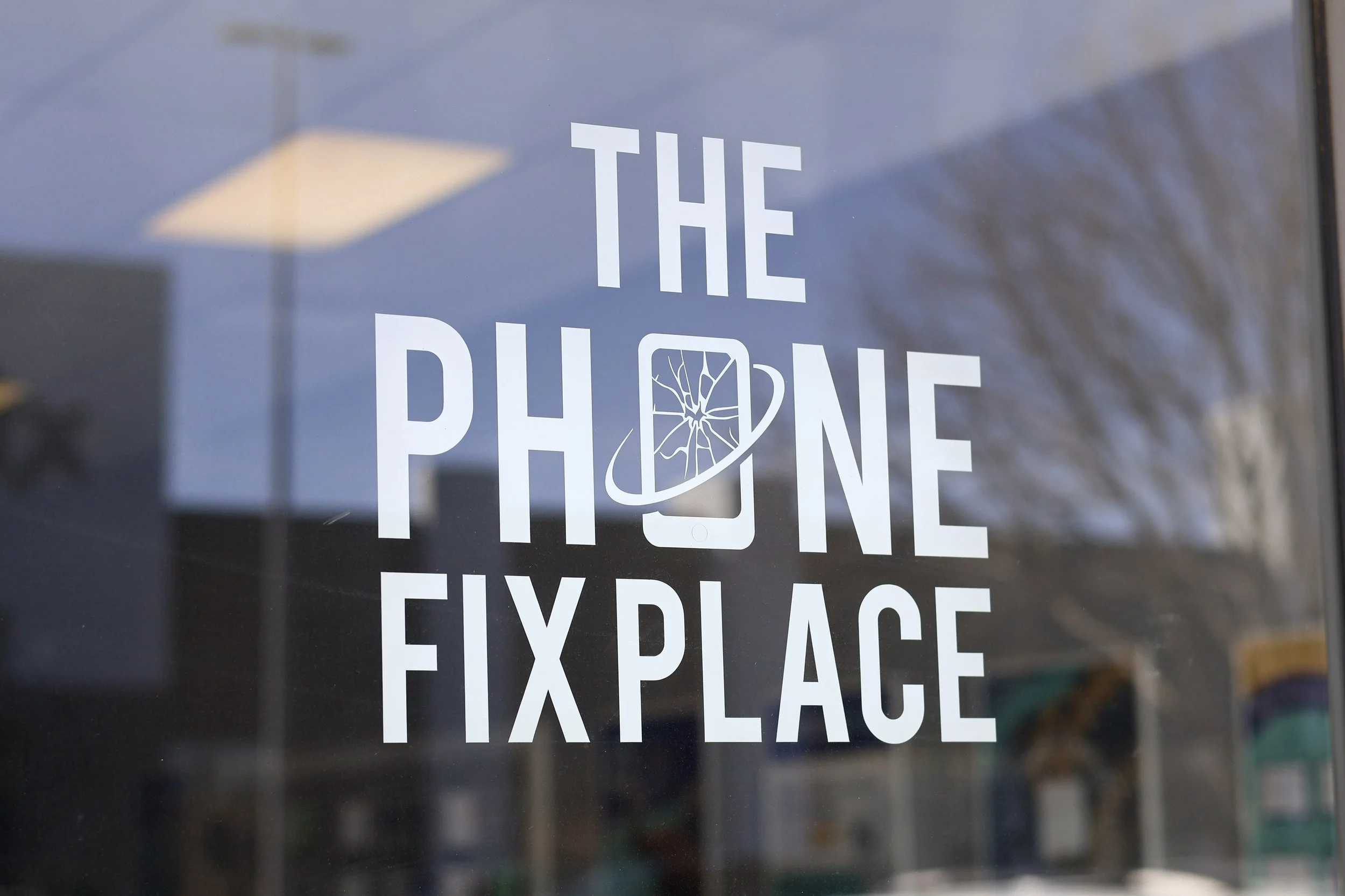 A window sign reads 'The Phone Fix Place' with a graphic of a smartphone featuring a cracked screen, and an orbiting ring around it.