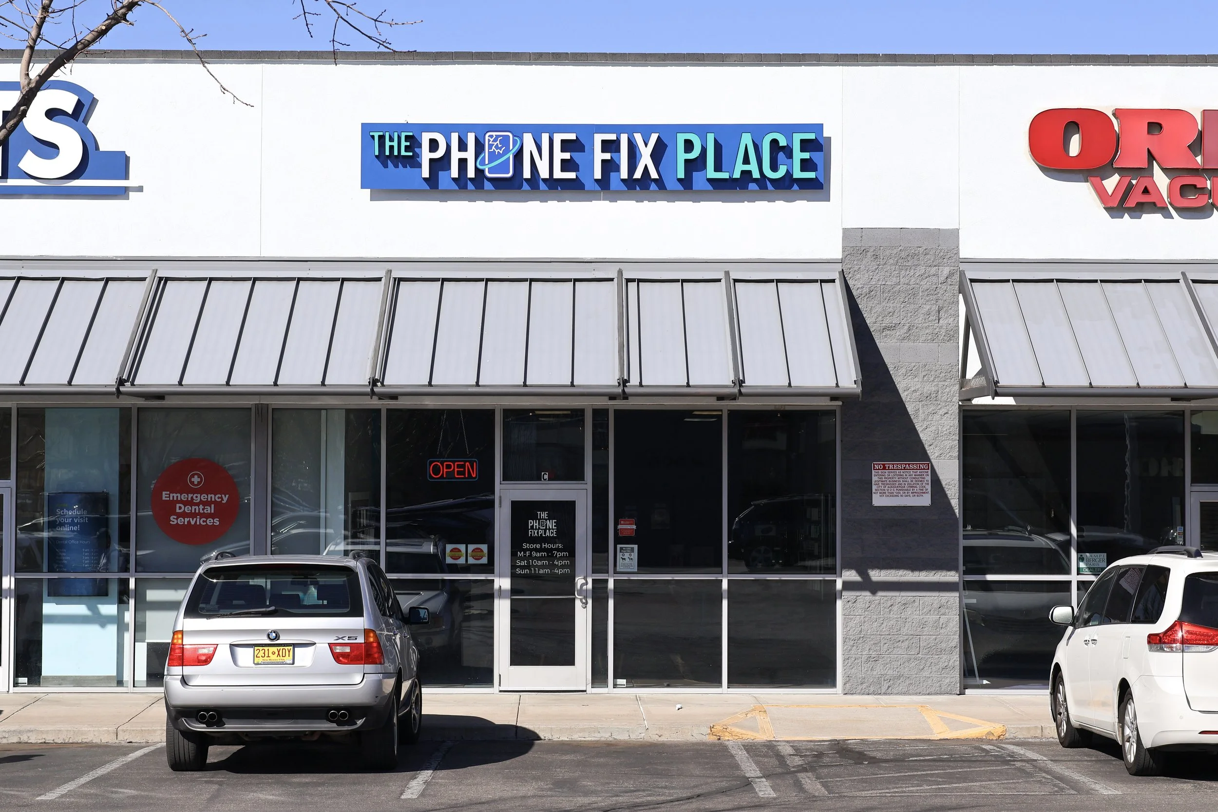 Store front of The Phone Fix Place with parked cars in front and signs indicating services like emergency dental and store hours.