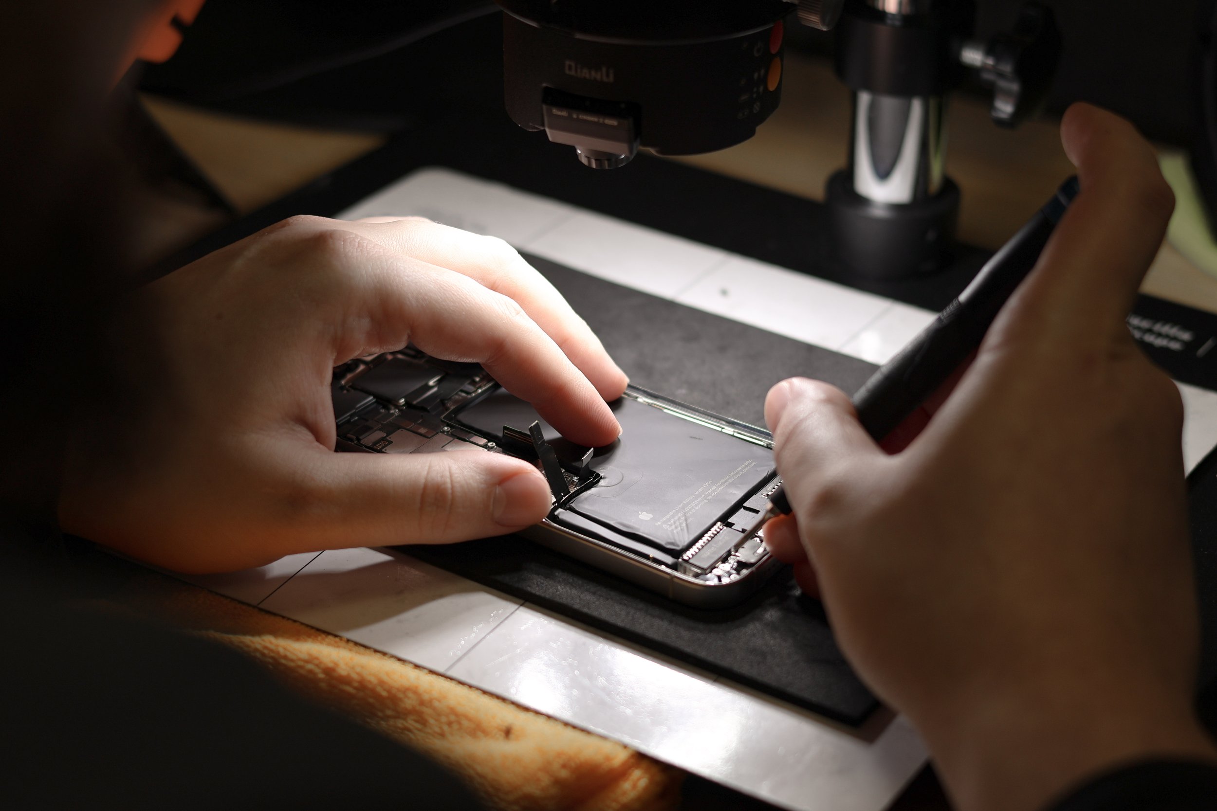 Person repairing a smartphone by inspecting its internal components with a tool under a microscope.