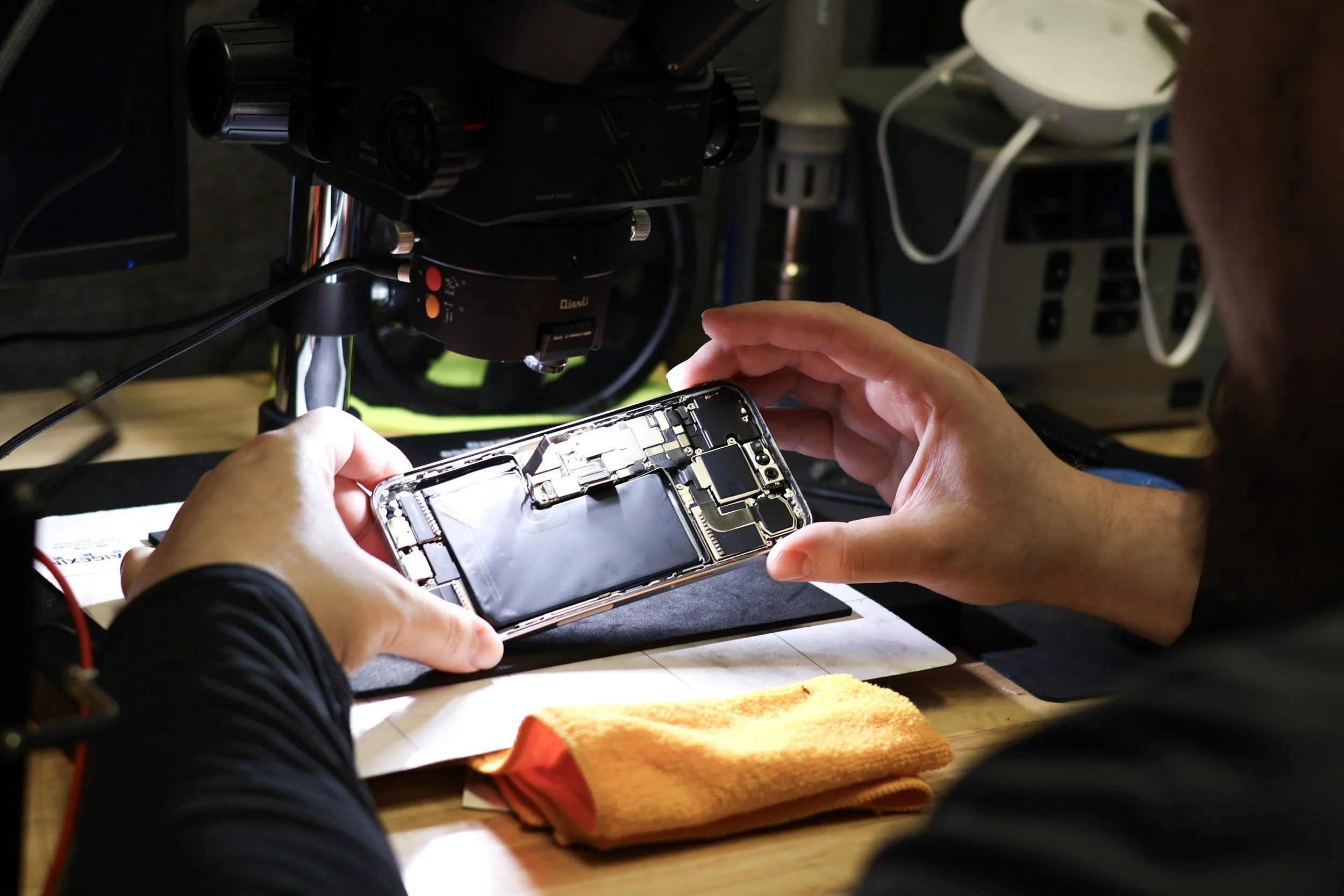 A person repairing a smartphone with an exposed interior, using a microscope or a similar device for close-up work, on a workbench with tools and a cloth.