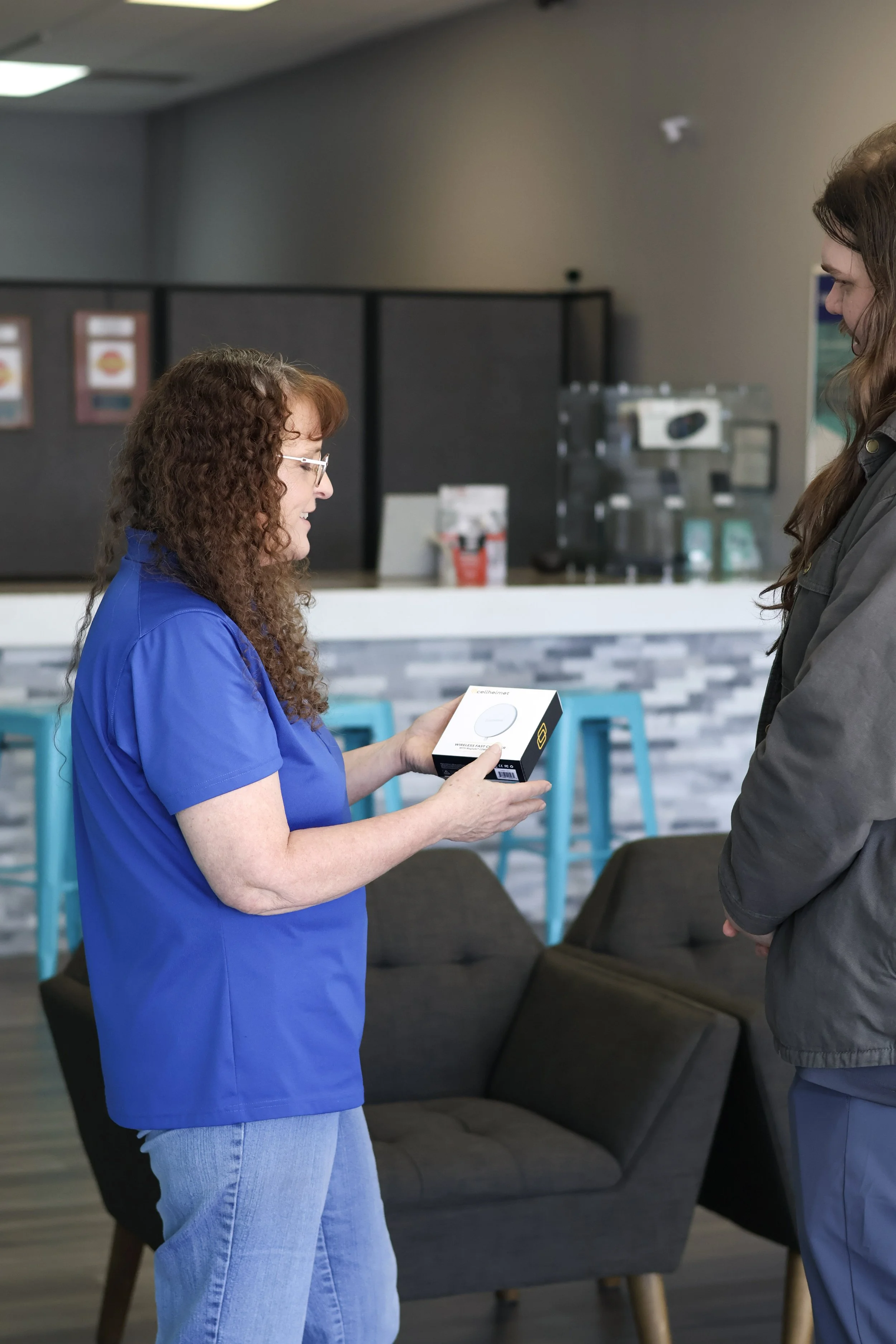 A woman in blue scrubs showing a box to another woman in a gray jacket inside a waiting room or lobby area.