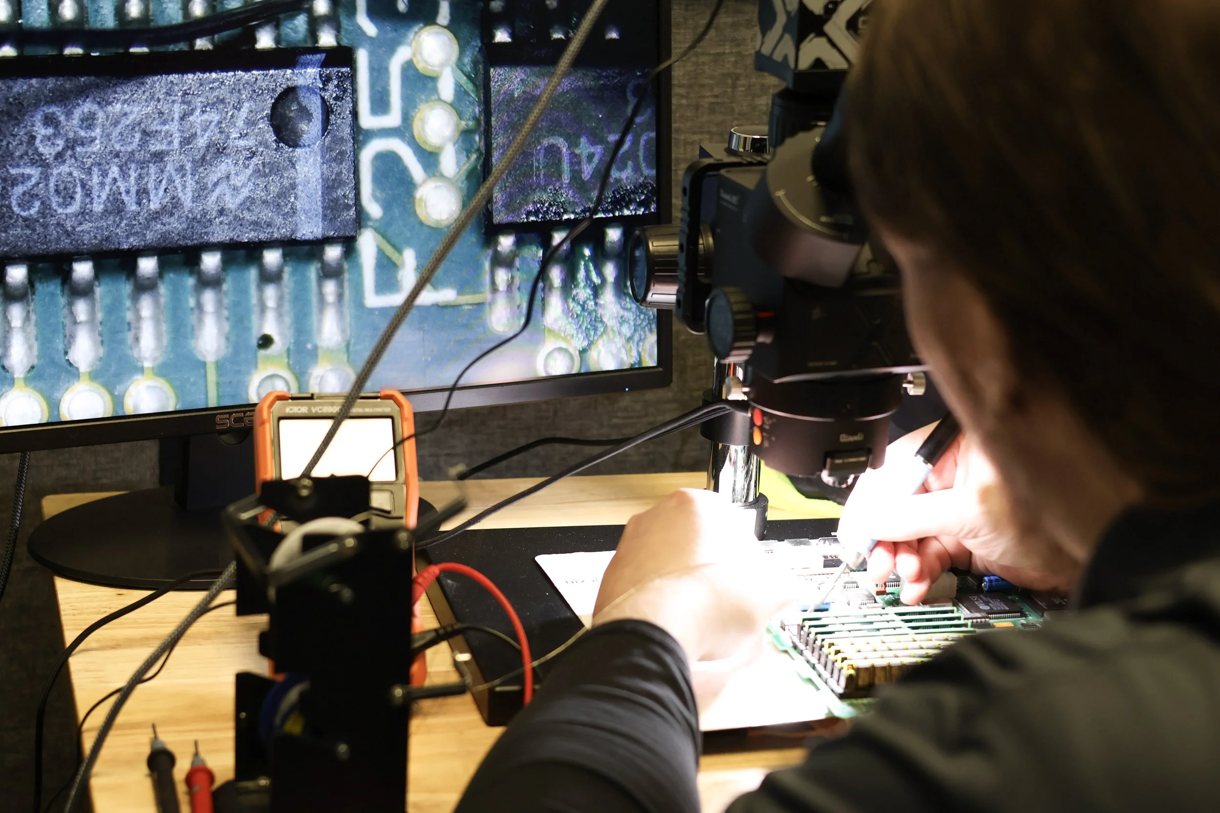 A person working with a microscope on an electronic circuit board, with a computer monitor displaying a close-up of the circuit.