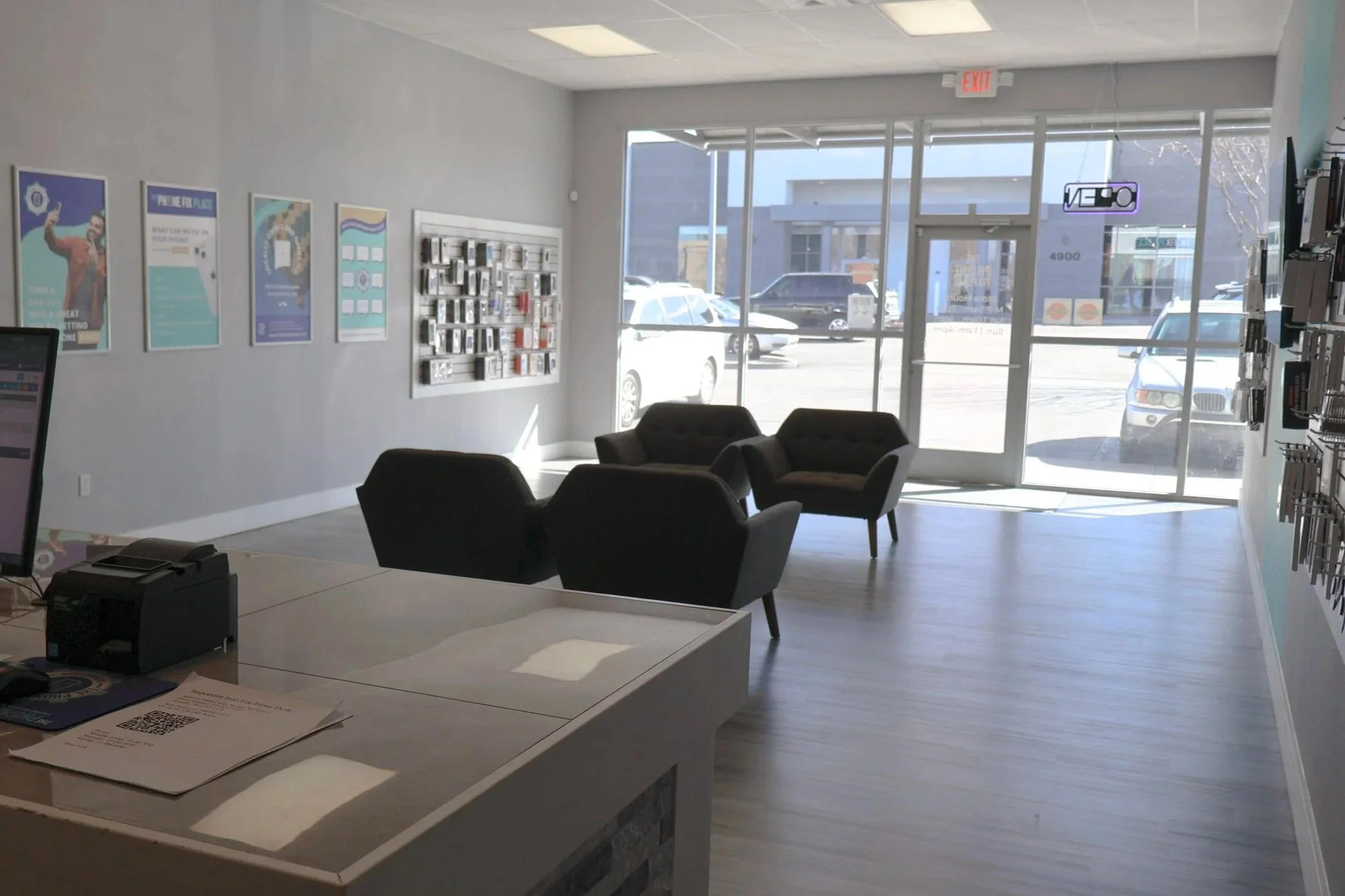 Empty waiting area with three black chairs in front of a glass door and large window, with sunlight coming in. Walls decorated with posters and a key rack, near a reception desk.