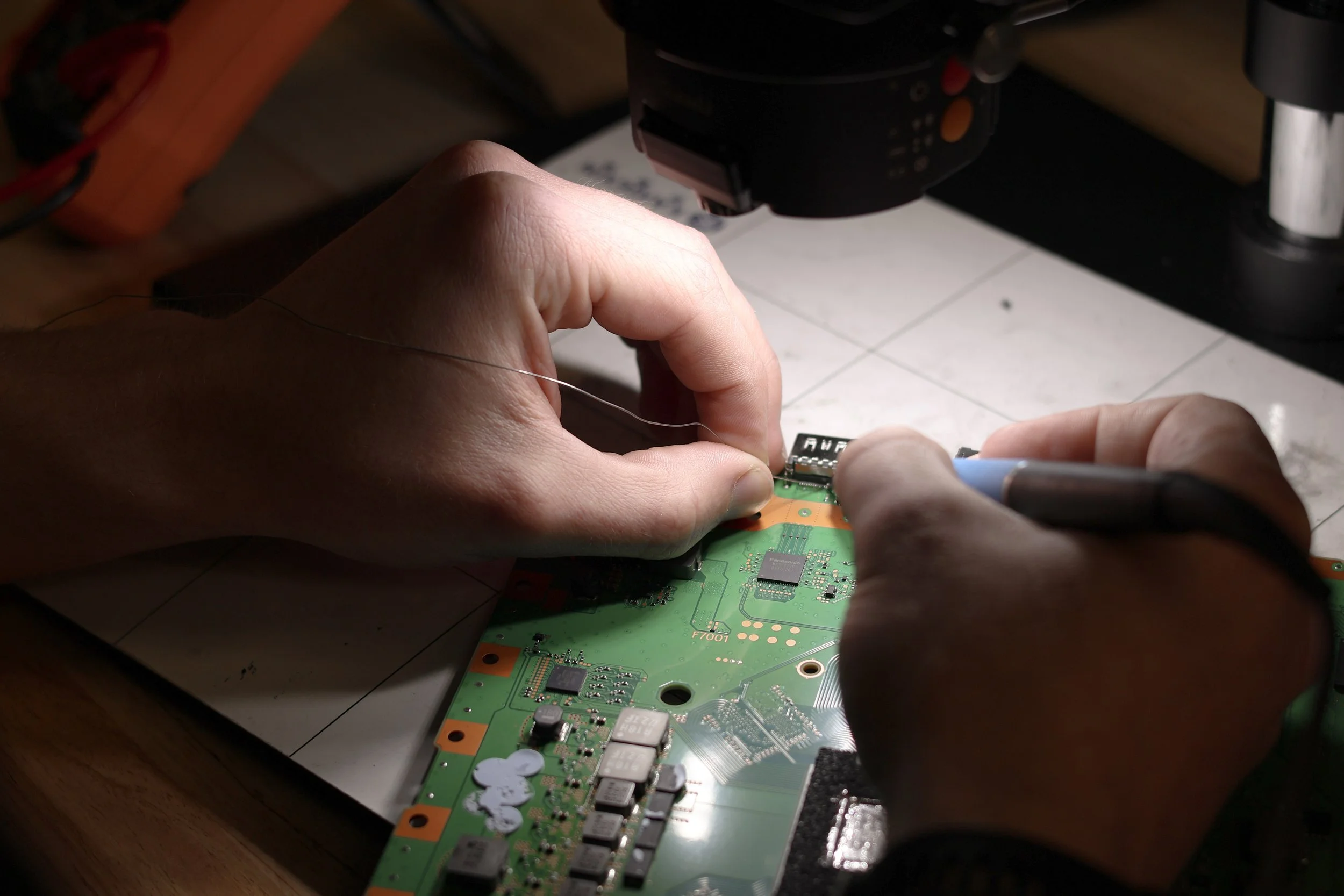 A person is repairing or assembling a printed circuit board (PCB) using a soldering iron under a microscope.