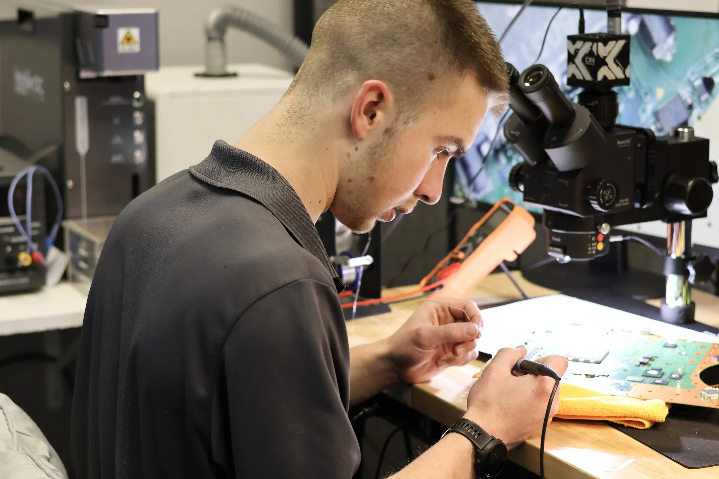 A young man in a black polo shirt working on an electronic circuit board at a workbench, with a microscope and various electronic equipment in the background.
