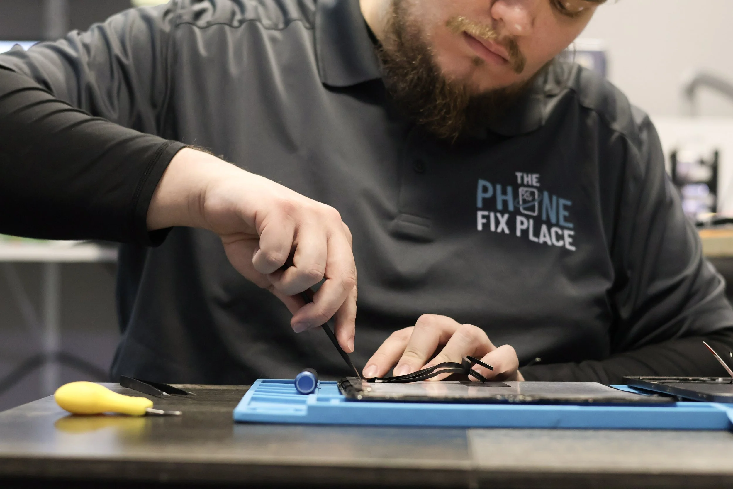 A technician wearing a gray shirt with a logo that says 'The Phone Fix Place' is repairing a smartphone with tools on a workbench.