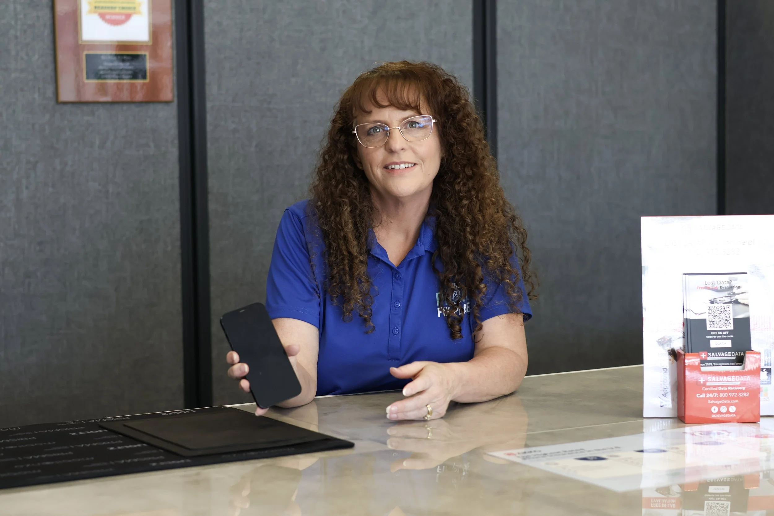 Woman with curly hair and glasses sitting at a reception desk, holding a smartphone in her right hand, in an office environment.