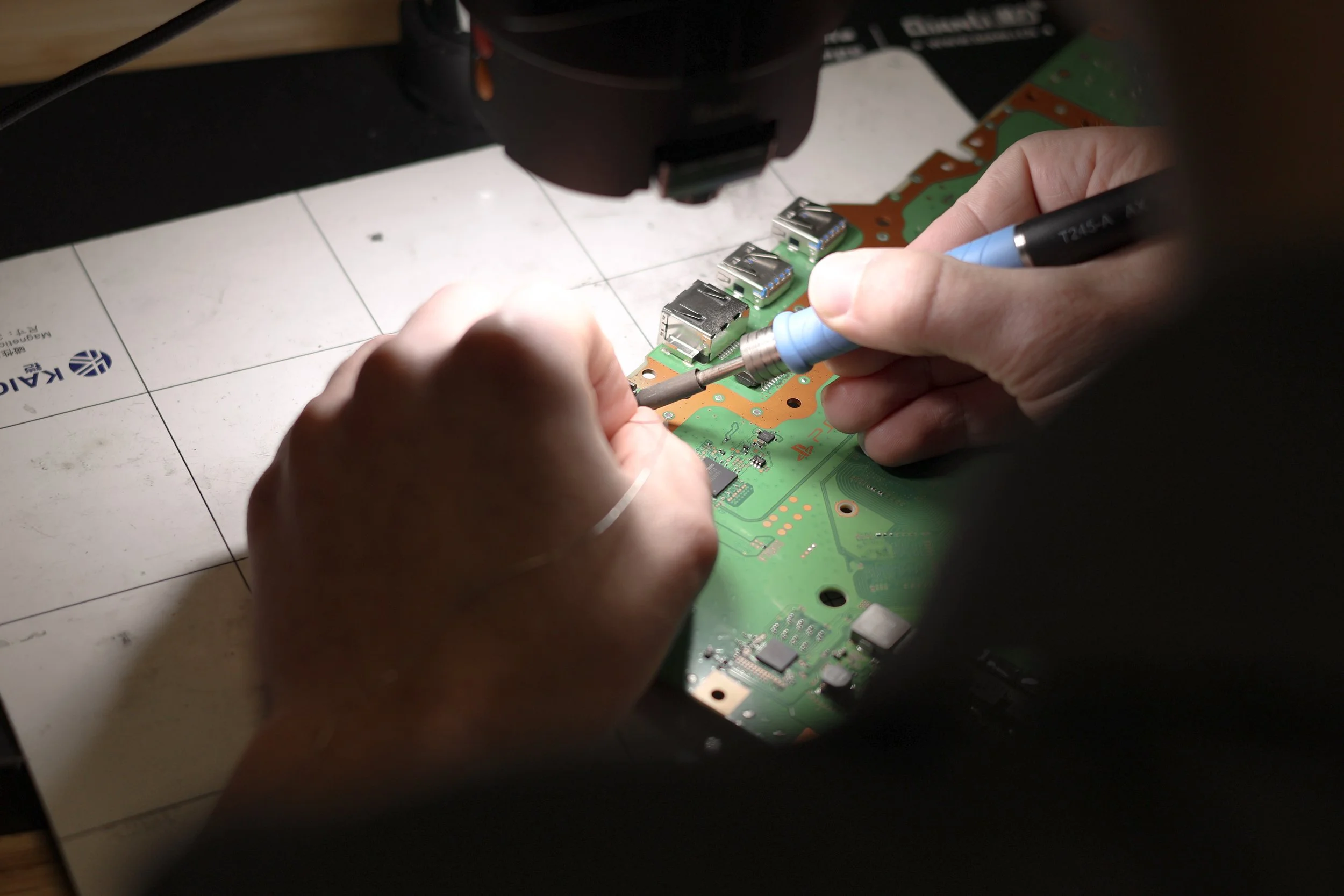 Person working on repairing a computer motherboard with a small screwdriver under a magnifying lamp.