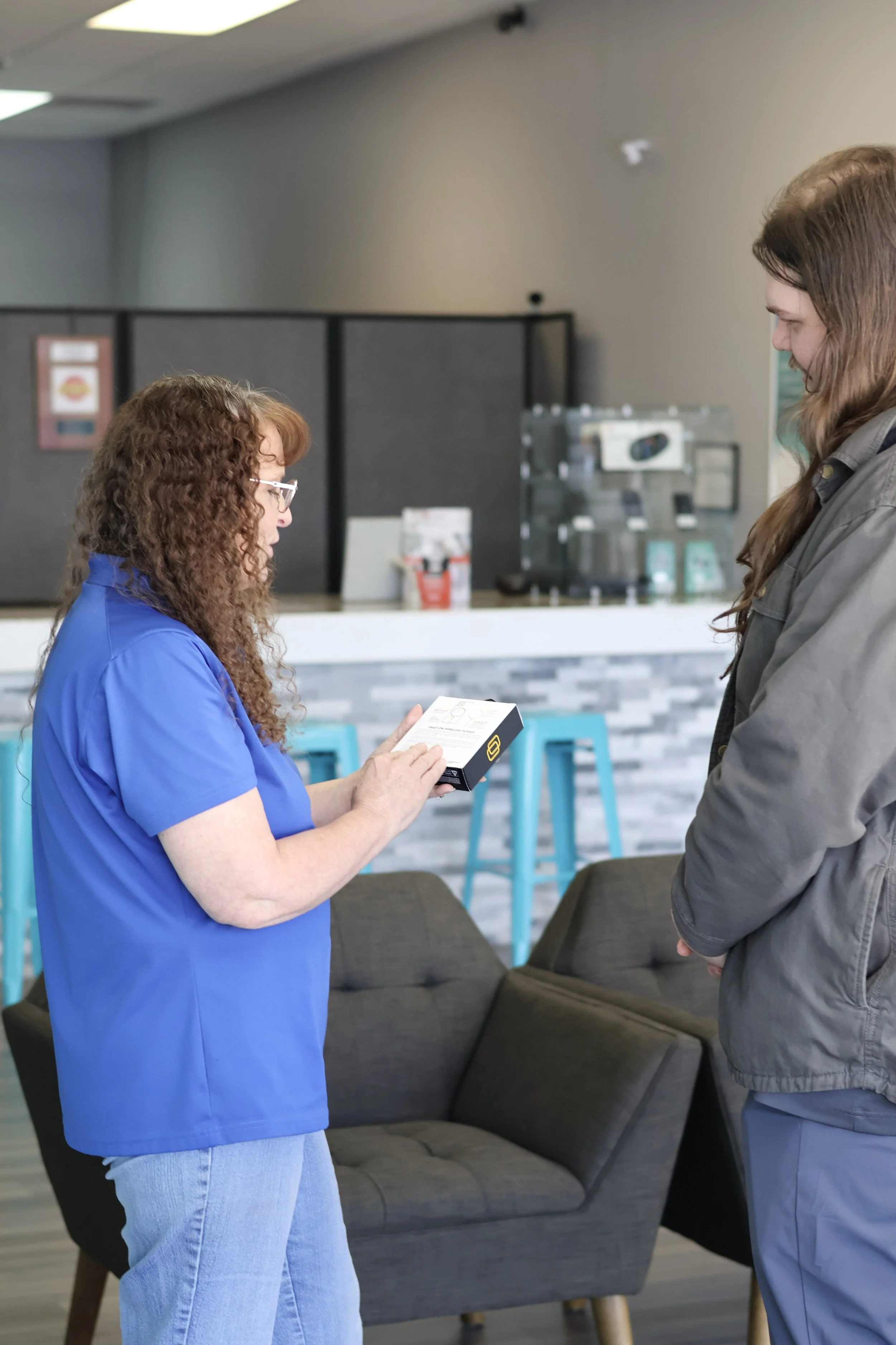 A woman in a blue shirt showing a small box to another woman in a gray jacket inside a modern cafe with bar stools and a counter in the background.