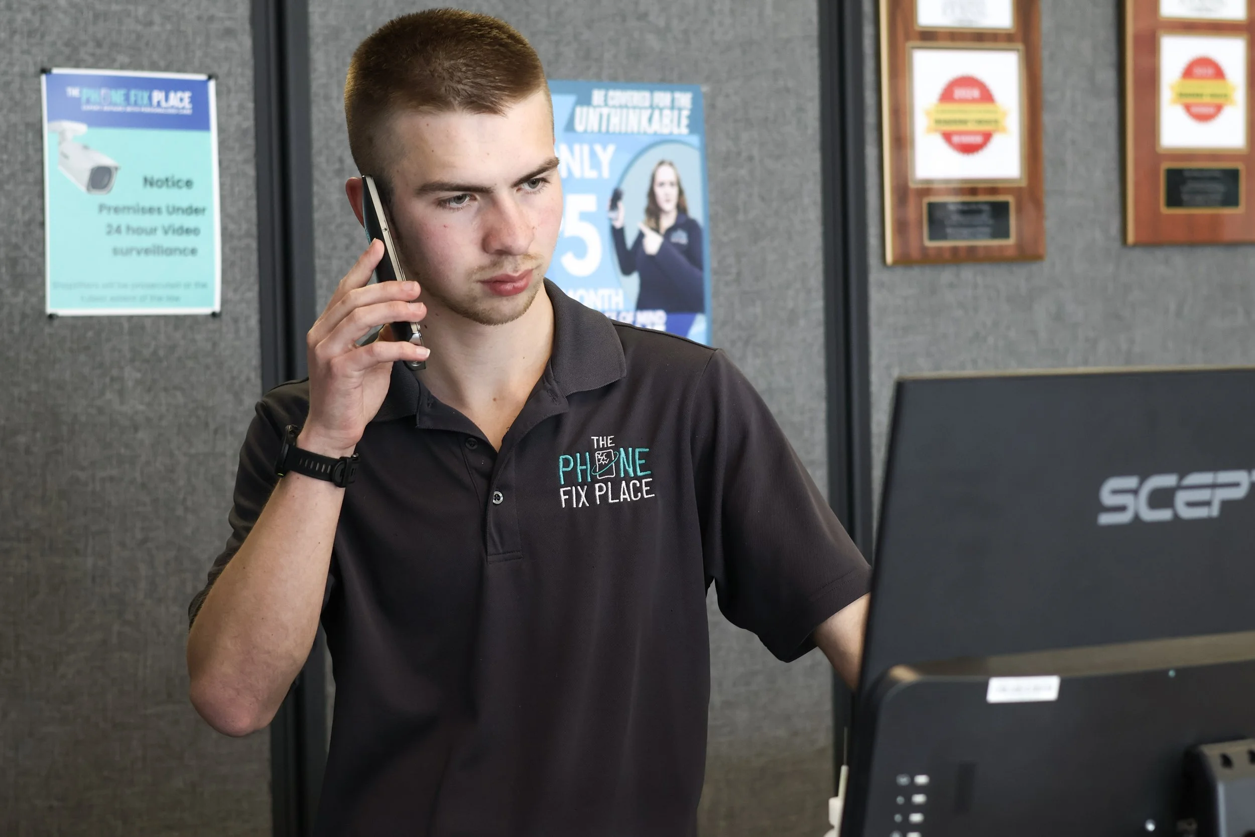 A young man with short brown hair and a black polo shirt with the logo 'The Phone Fix Place,' standing at a counter with a computer, talking on a cell phone inside a tech repair shop.