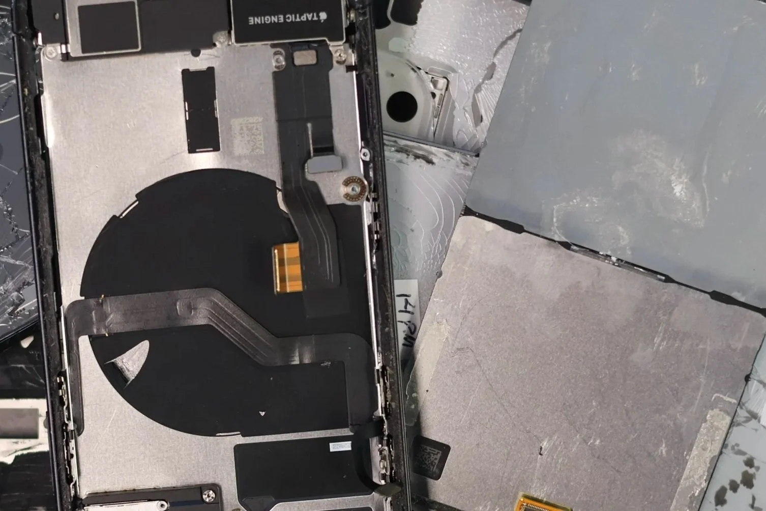 Interior view of a damaged electronic device, showing circuit boards, ribbon cables, and a circular component inside a metal frame with some debris and damage visible.