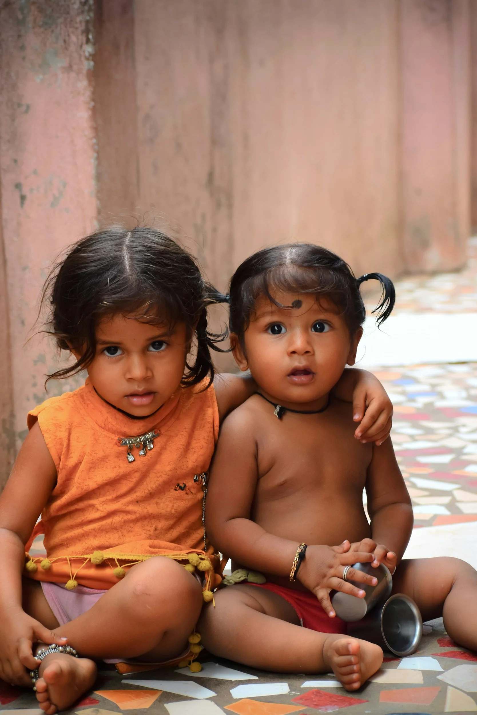 Two young children, a girl and a boy, sitting on a patterned floor against a pinkish wall, with the girl having her arm around the boy. The girl has dark hair styled with pigtails, wearing an orange top and jewelry. The boy is shirtless, wearing red shorts, with short dark hair and jewelry, holding a metallic cup.
