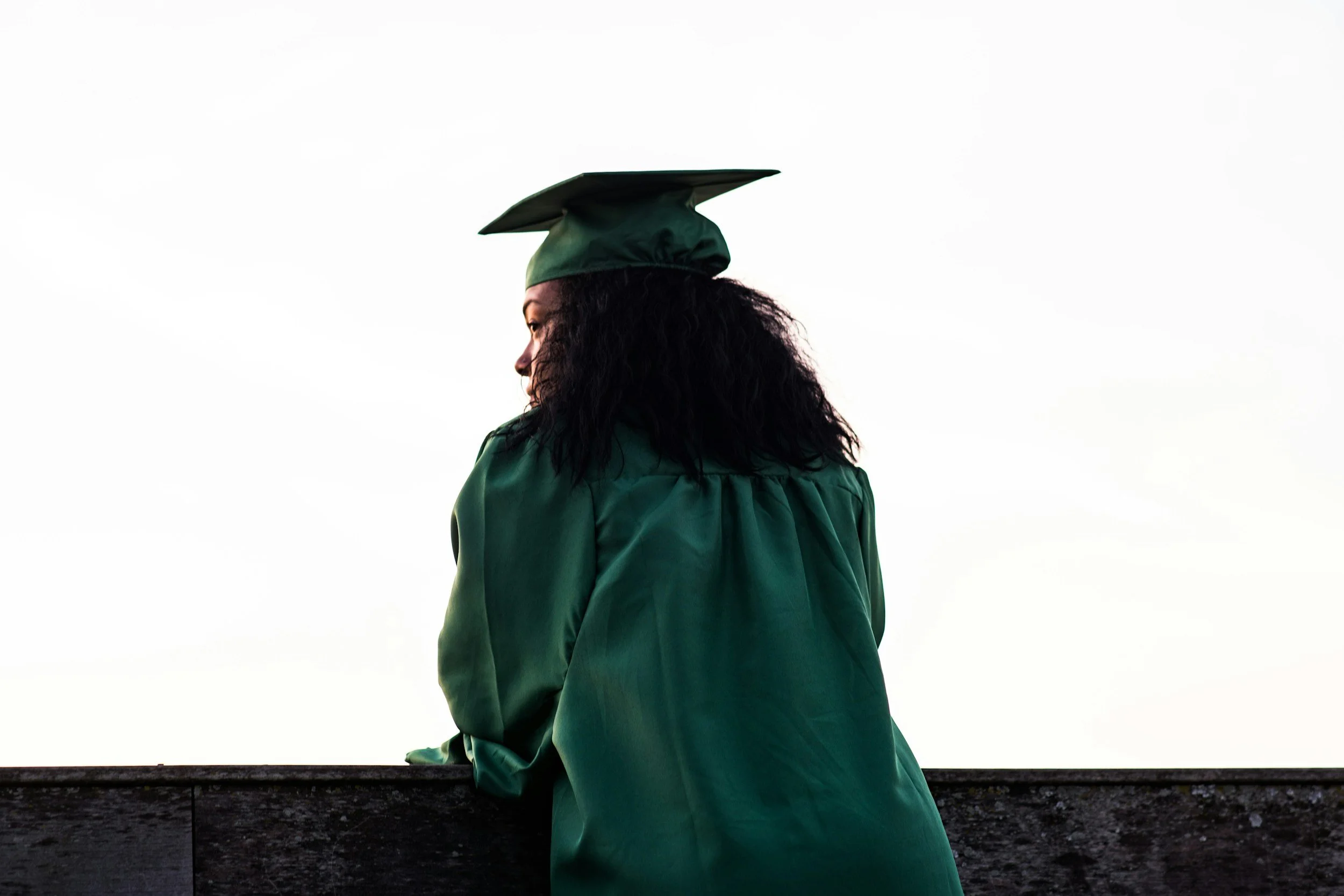 A woman in a green graduation gown and cap leaning on a railing, looking to the side, against a bright sky.