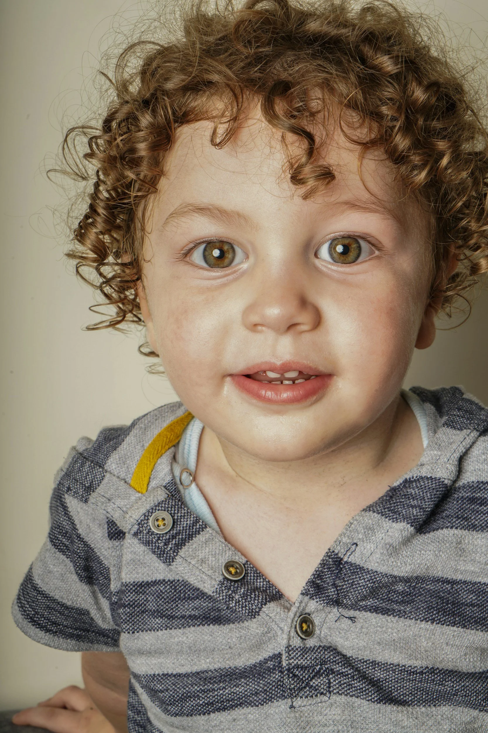 Close-up of a young boy with curly brown hair and hazel eyes, smiling slightly, wearing a grey and navy striped shirt with buttons.