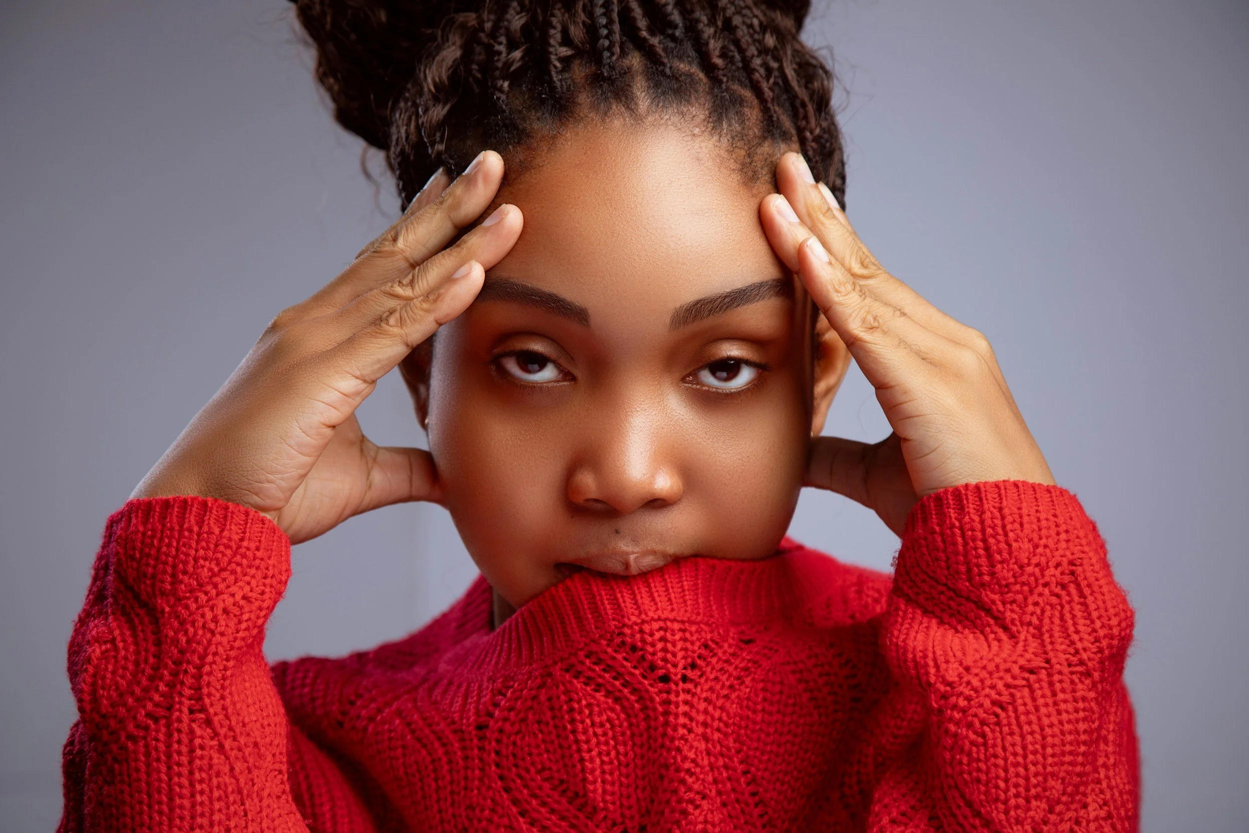 A woman with braided hair wearing a red sweater, holding her head with her hands, looking directly at the camera with a serious expression.