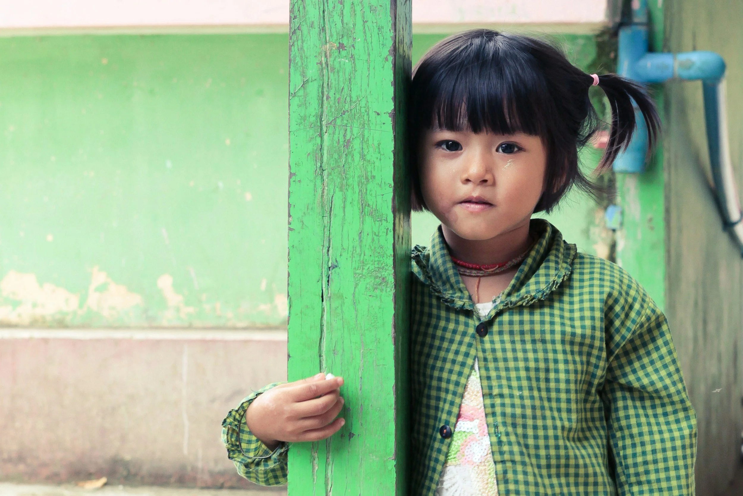 A young girl with dark hair tied in a ponytail peeking from behind a worn green wooden post in an indoor setting.