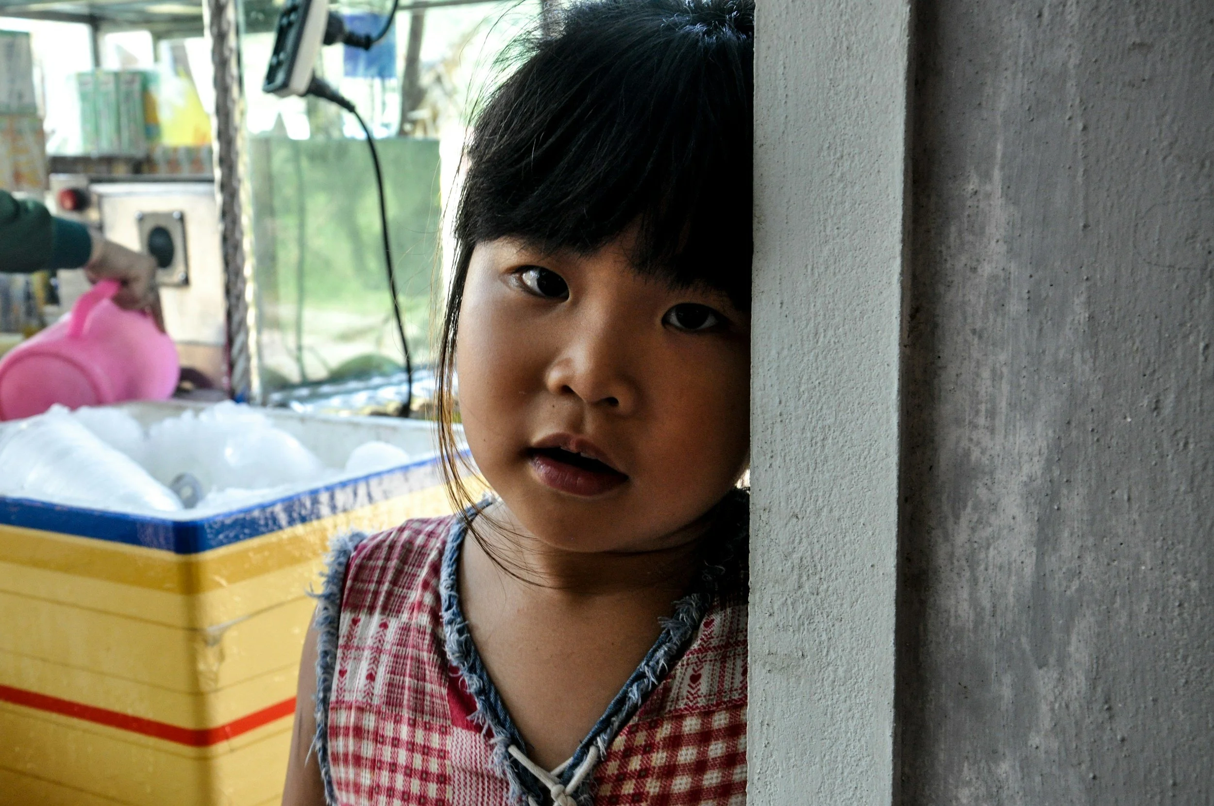 A young girl with black hair and a checkered shirt peeking from behind a wall, looking at the camera.
