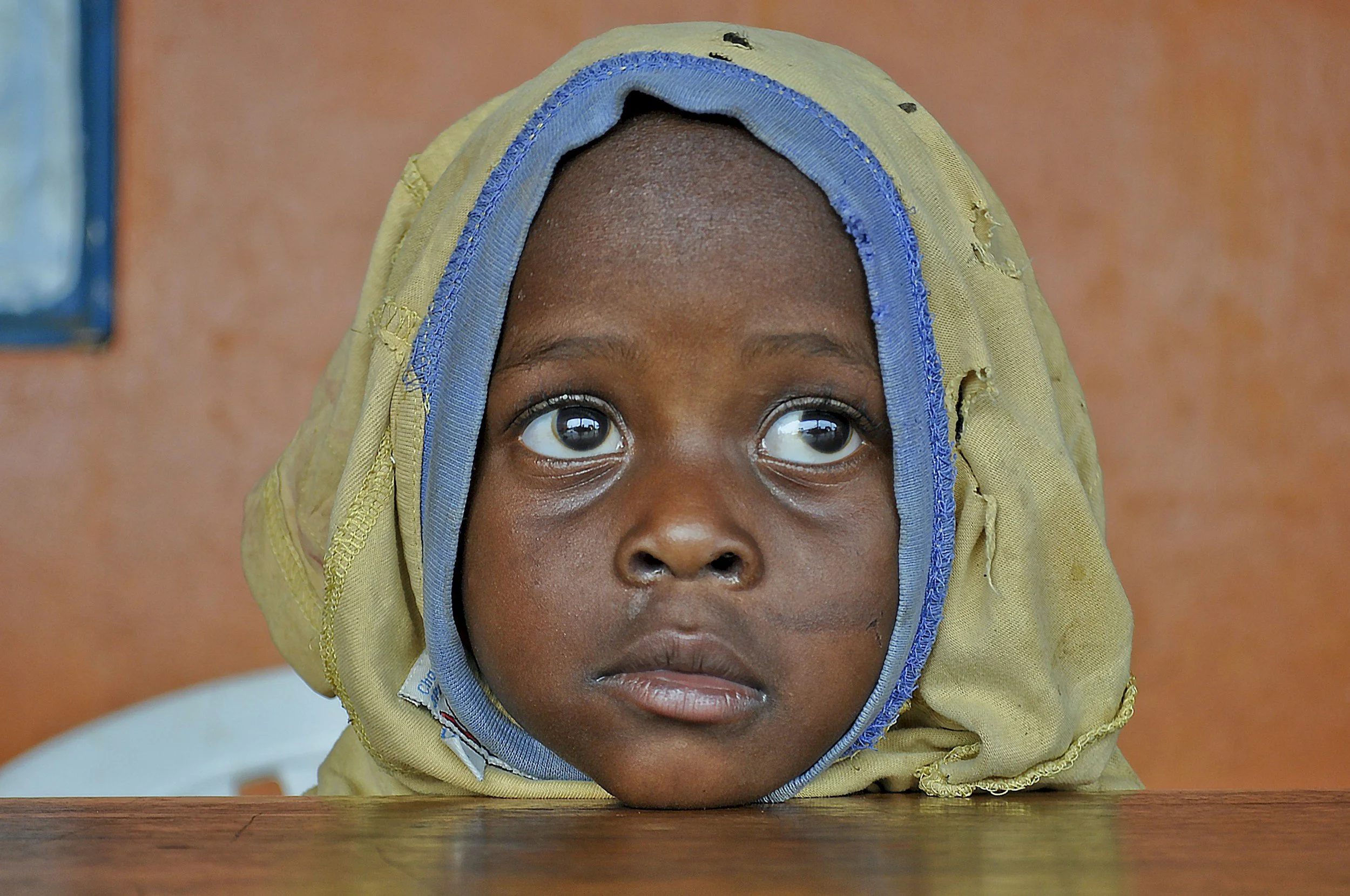 Close-up of a young girl with a hooded head cover, looking upward with wide eyes, seated at a wooden table.