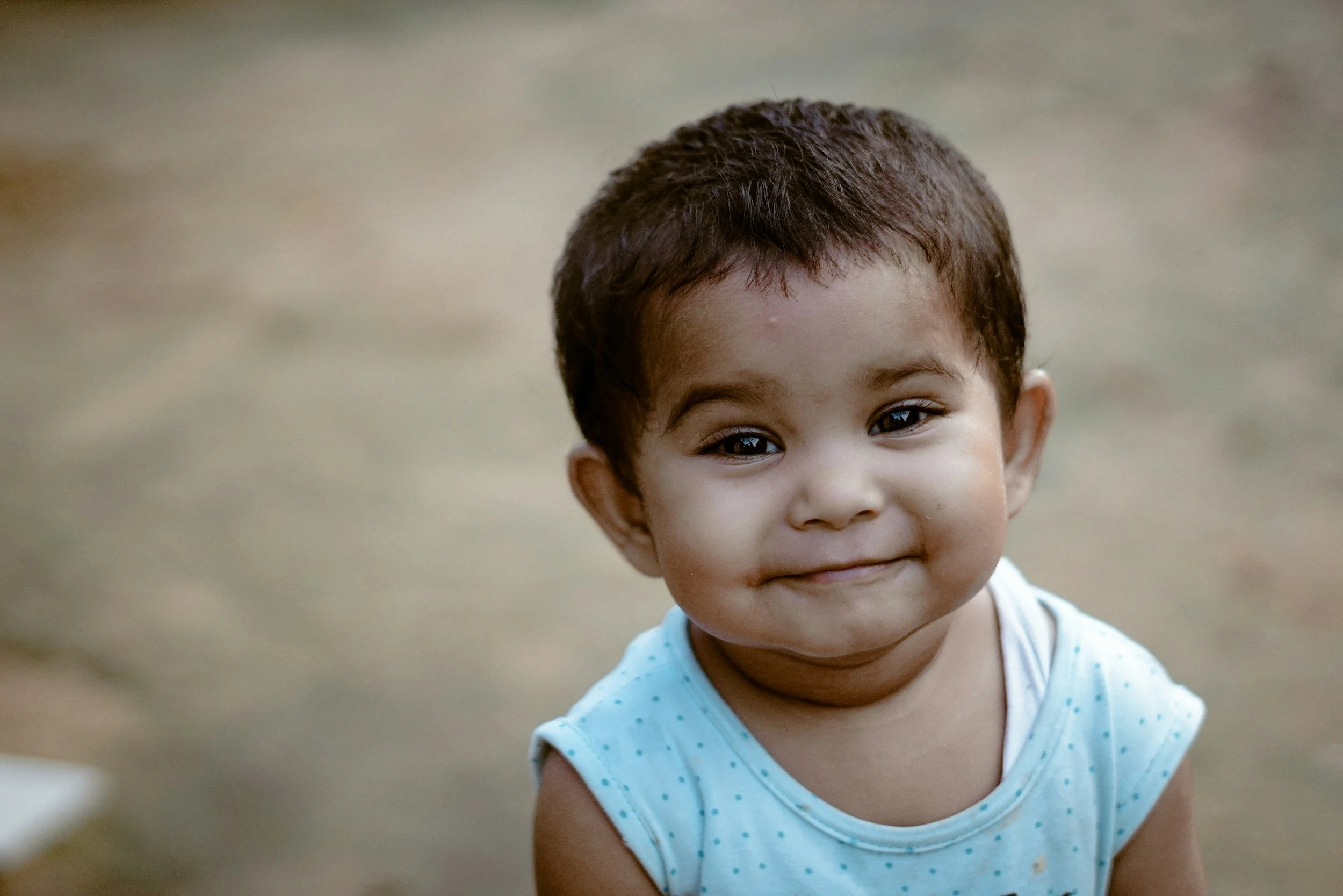 Close-up of a young child with short dark hair, smiling softly, wearing a light blue sleeveless shirt.