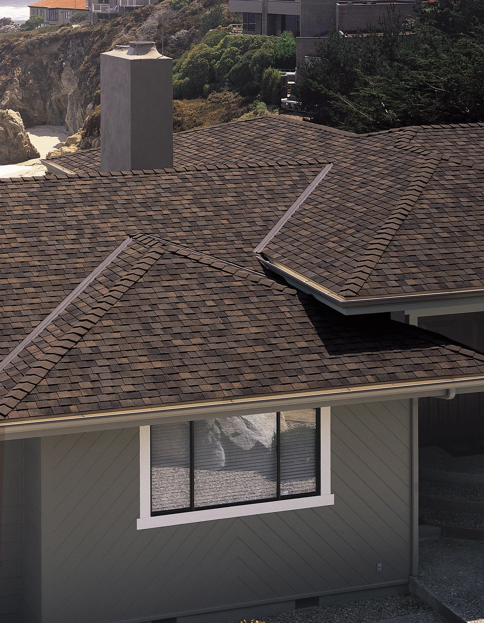 View of a house rooftop with brown shingles, a chimney, and a satellite dish. The house has large windows and grey siding, with trees and other houses in the background.